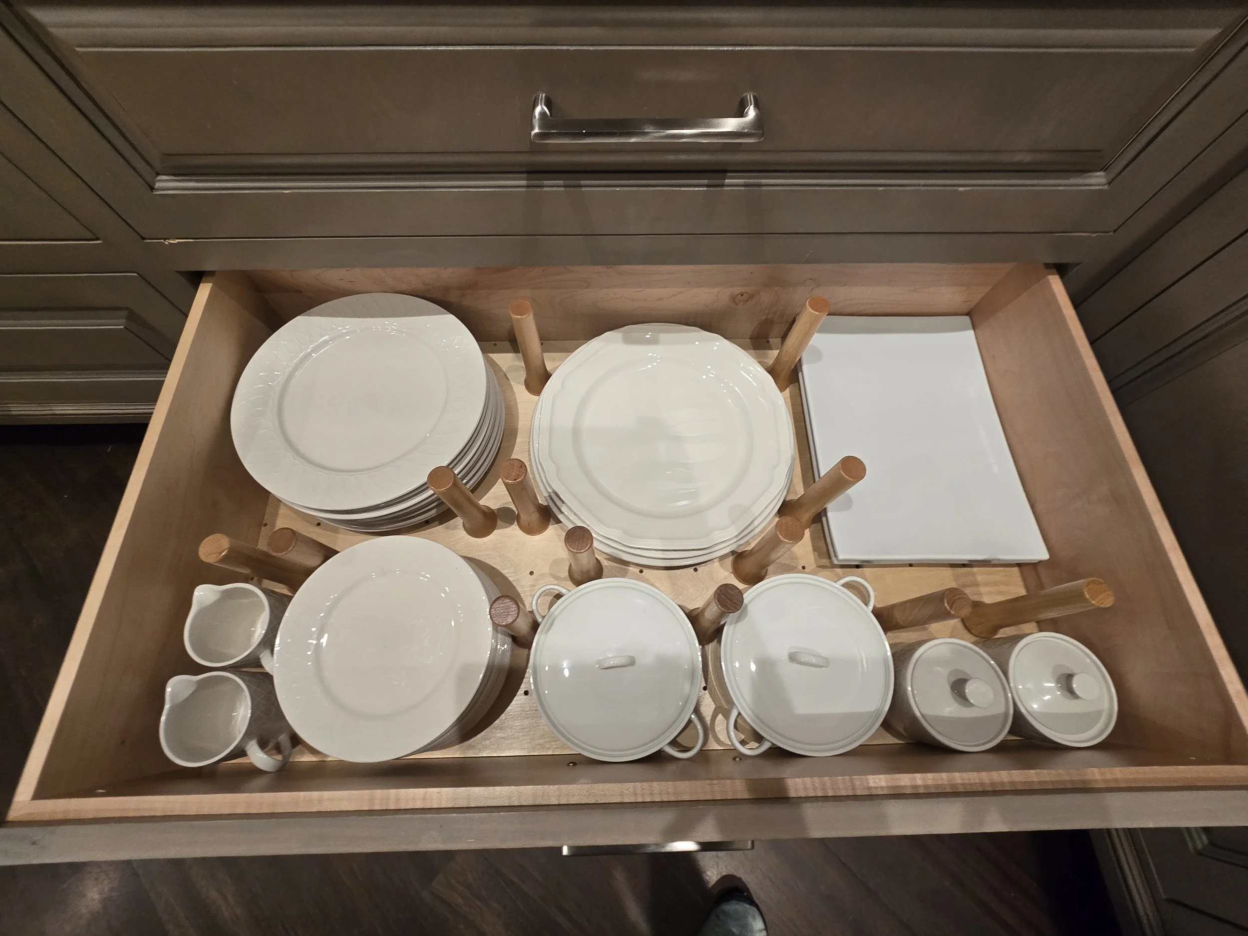 An open drawer with white plates, bowls, and mugs stacked and stored upright in a wooden kitchen drawer.