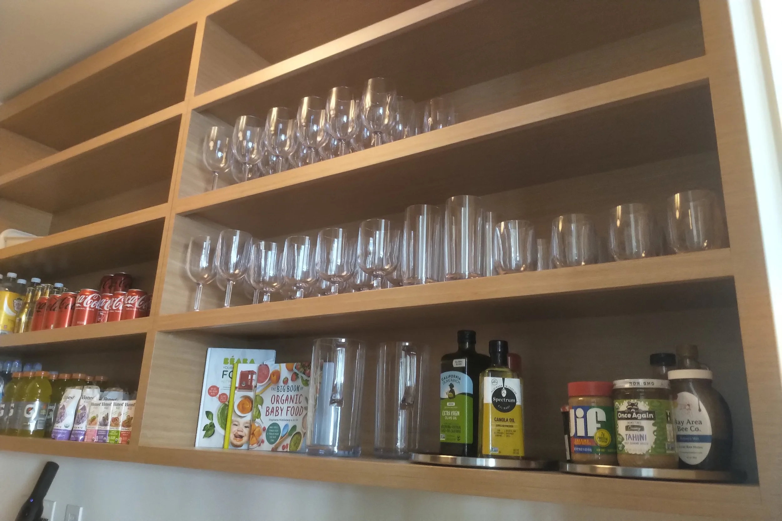 Open wooden kitchen shelf containing wine glasses, water glasses, canned drinks, cookbooks, oil bottles, and food jars.