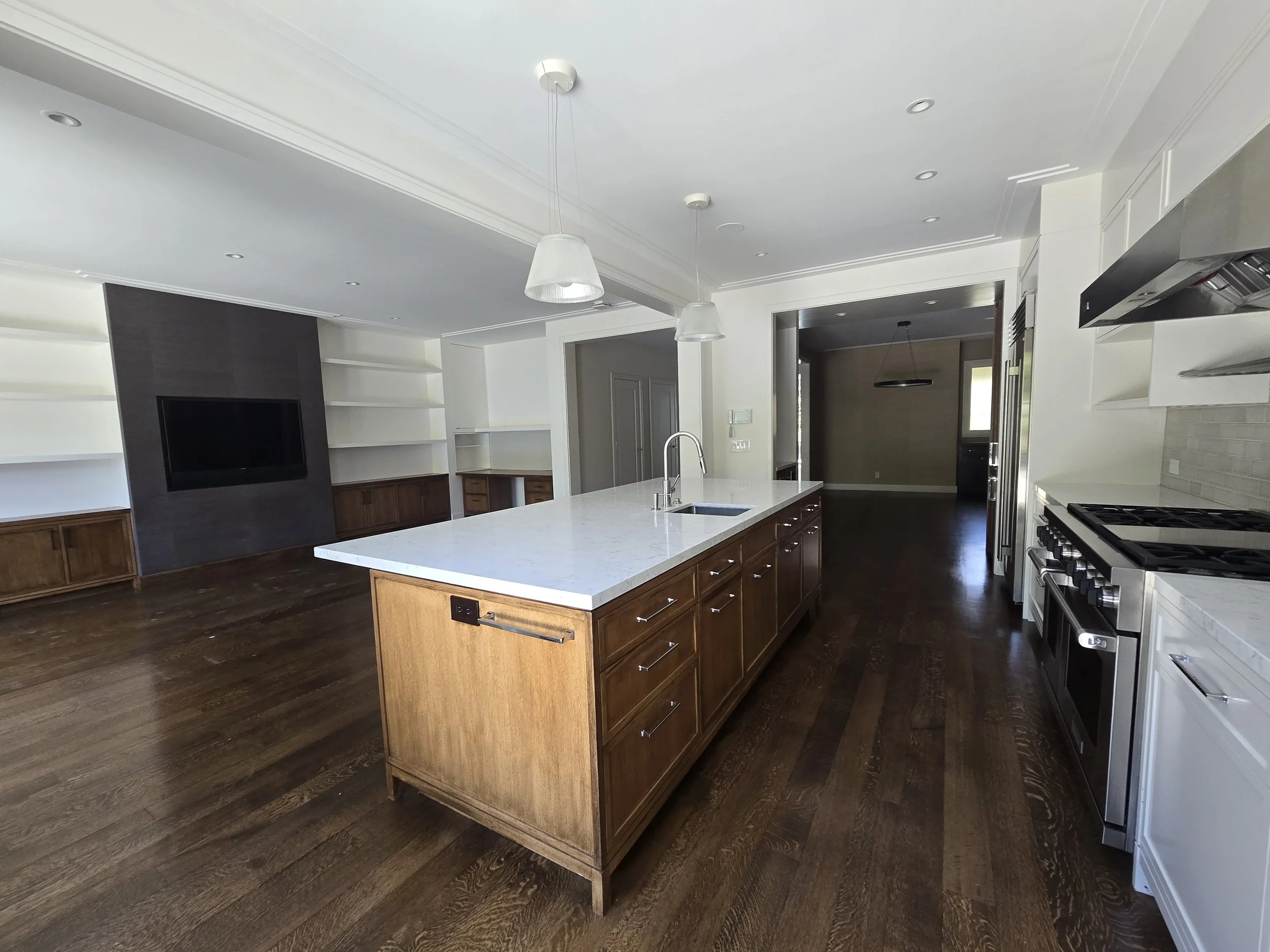 Modern kitchen with large island featuring white marble countertop, brown wooden cabinets, stainless steel appliances including oven and range, pendant lights above island, dark hardwood flooring, white walls, and open living space with built-in shel