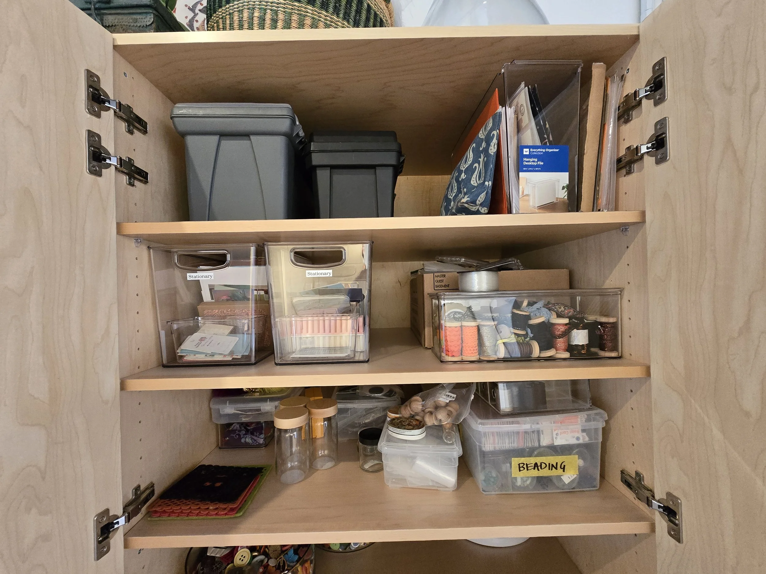 A wooden cabinet door opened to reveal three shelves filled with various office and craft supplies, including storage boxes, spools of thread, and labeled containers.