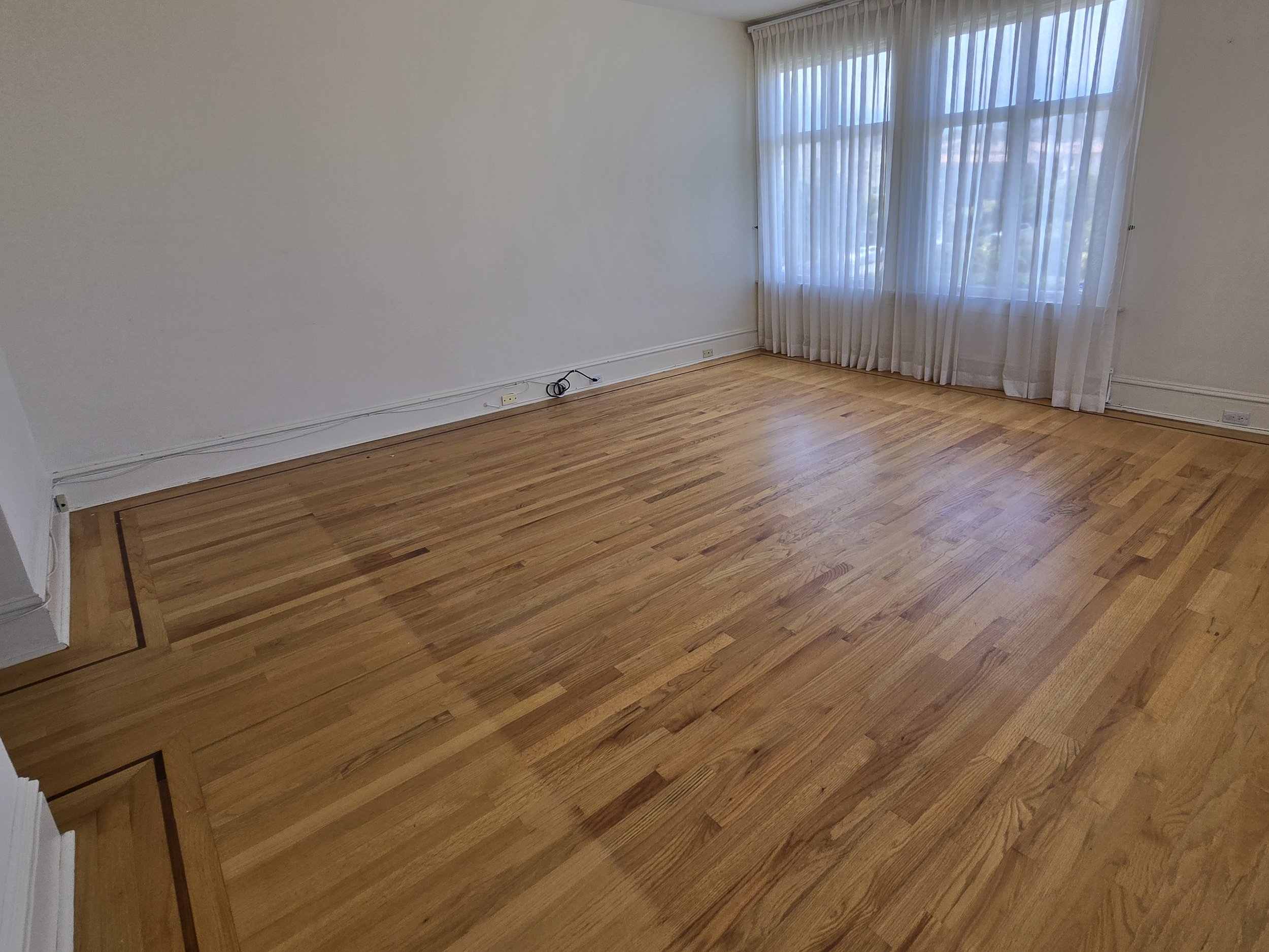 Empty room with wooden floor, white walls, and large window with sheer white curtains letting in natural light.