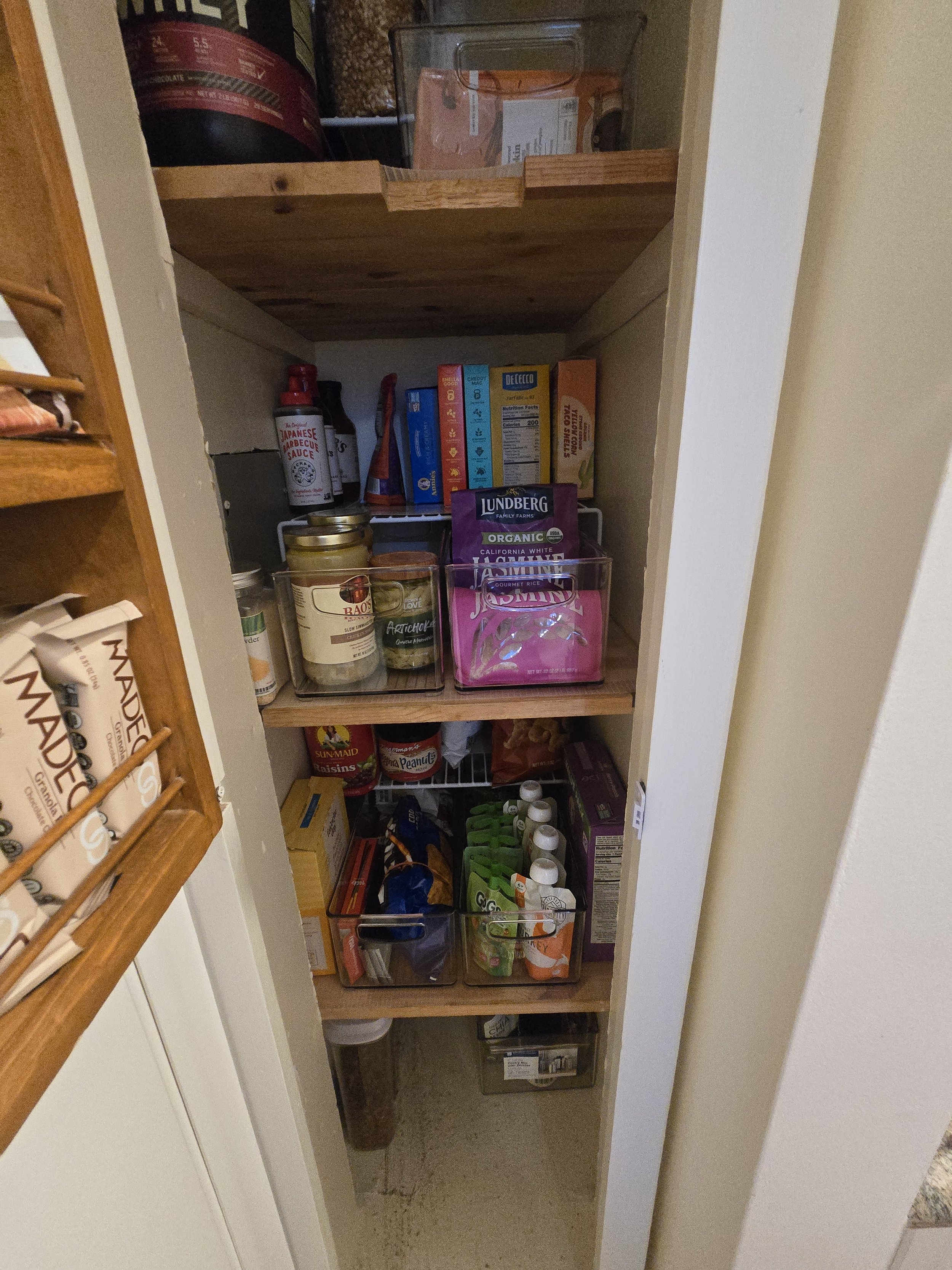 Pantry with wooden shelves holding various food items, spices, snacks, beverages, and storage containers.