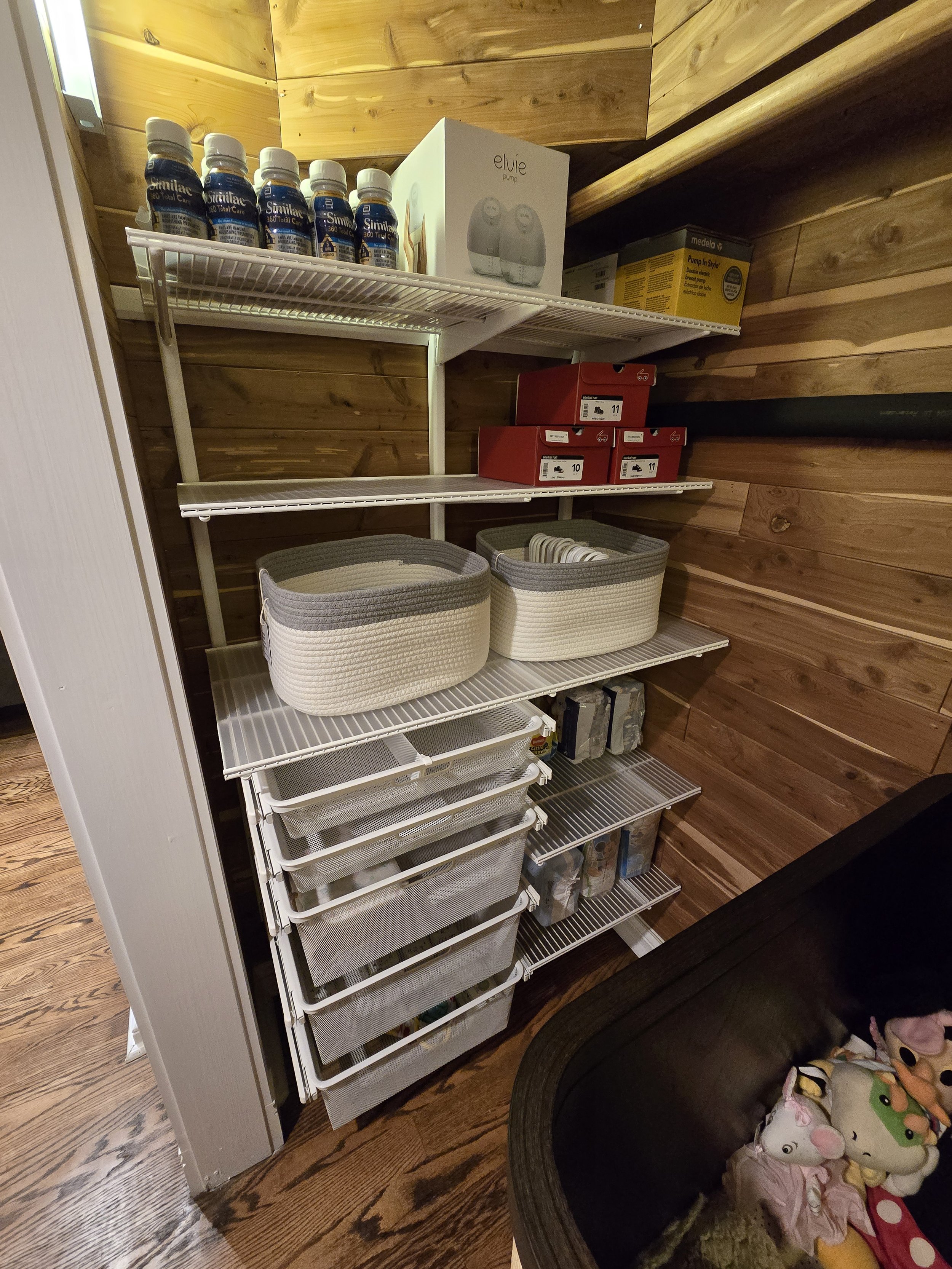 A closet with wooden walls and white wire shelving holding bottles, boxes, baskets, and containers, with plush toys in a black container at the bottom right.