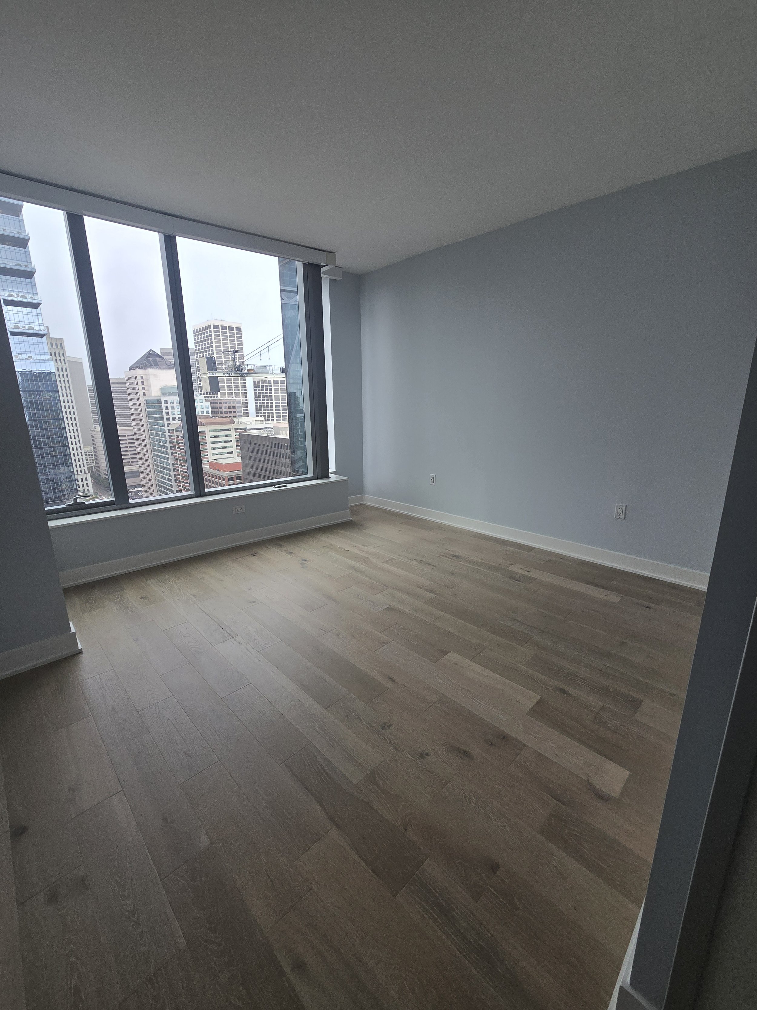 Empty room with large floor-to-ceiling windows overlooking a city skyline of tall buildings.