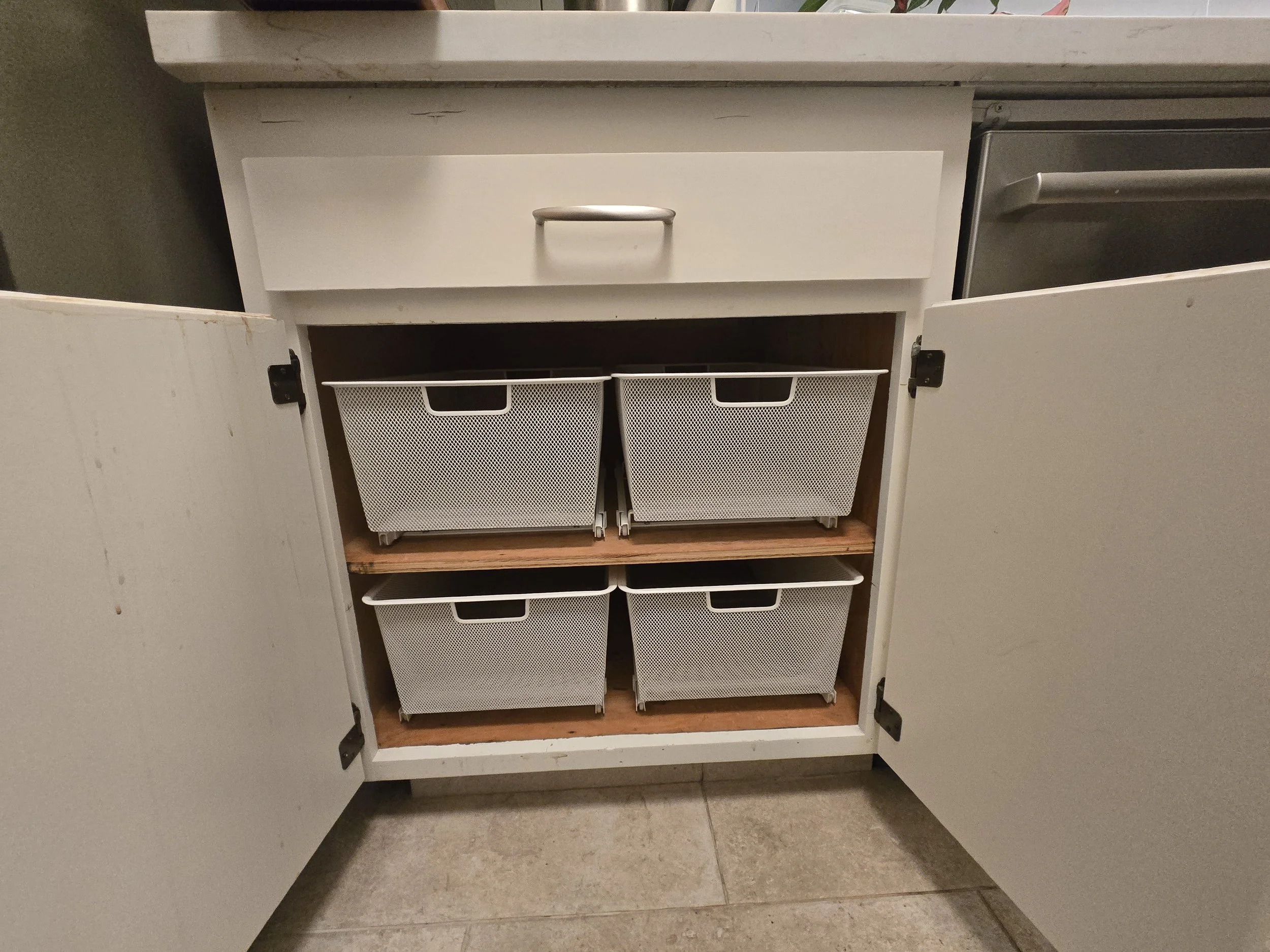 Open kitchen cabinet with four white mesh storage bins inside, two on each shelf, in a tiled kitchen floor.