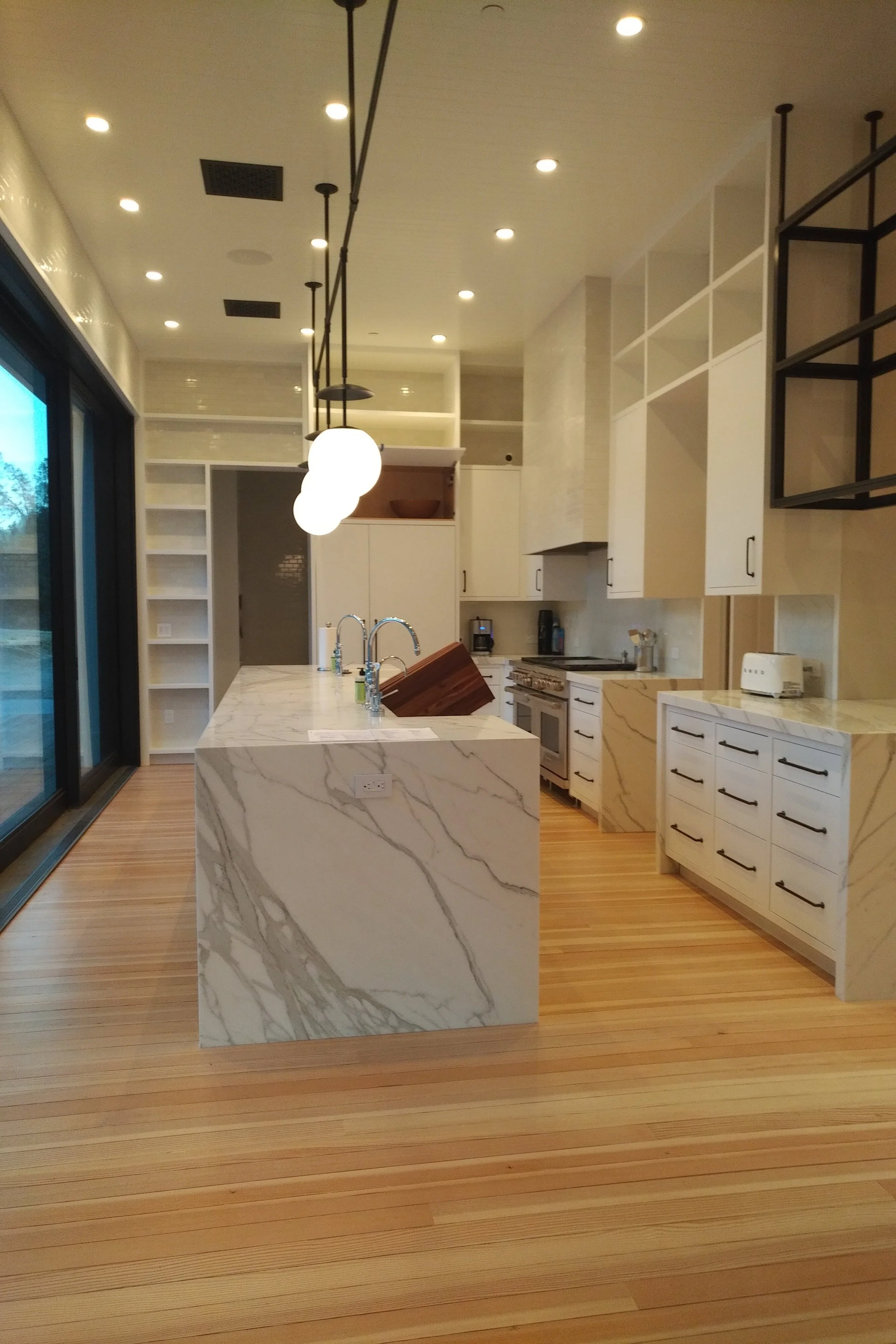 Modern kitchen with white cabinets, marble island, wooden flooring, black pendant lights, and large sliding glass doors.