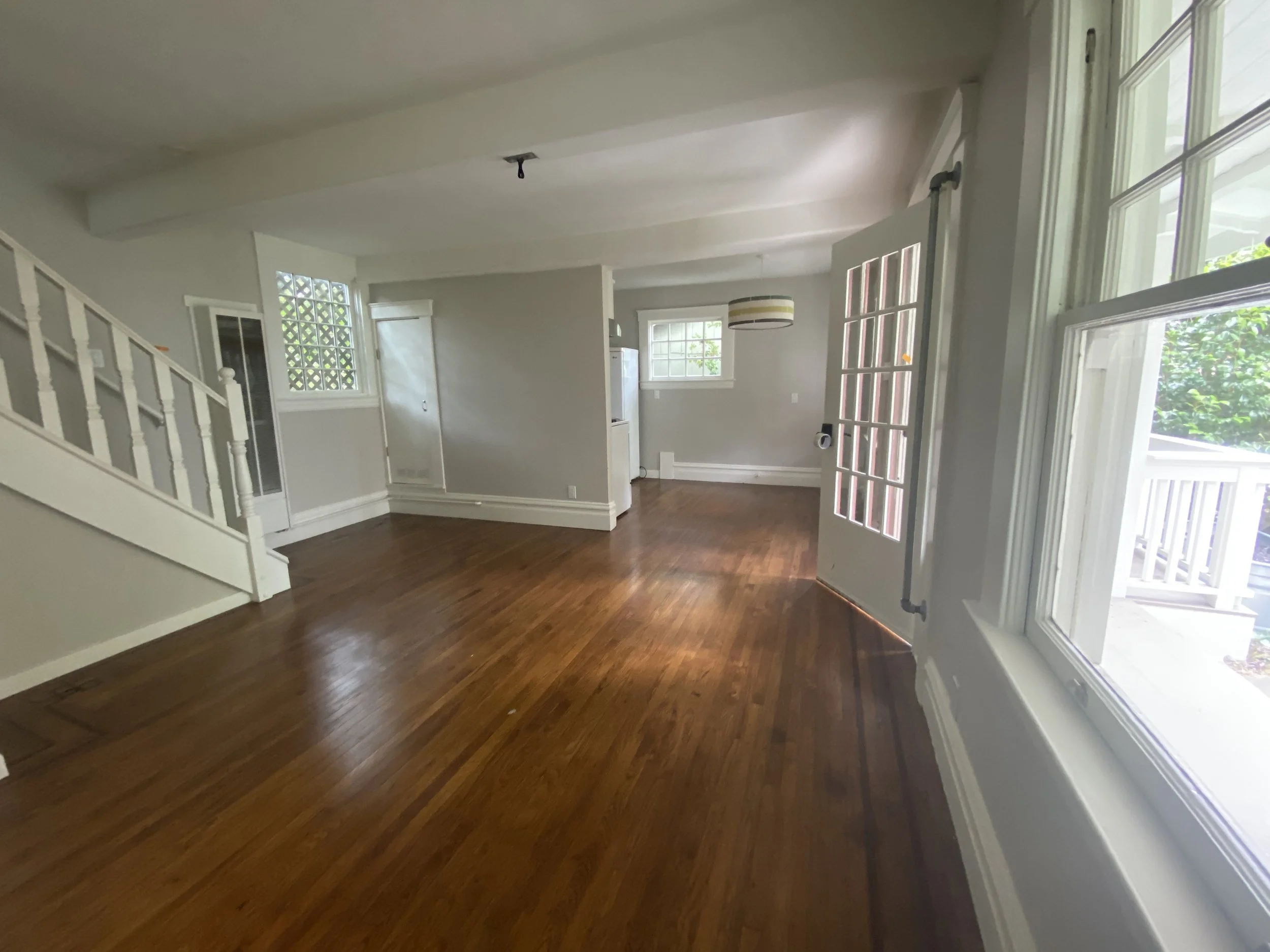 Empty living room with hardwood floors, white walls, multiple windows, a staircase on the left, and a partially open door leading to the outside on the right.