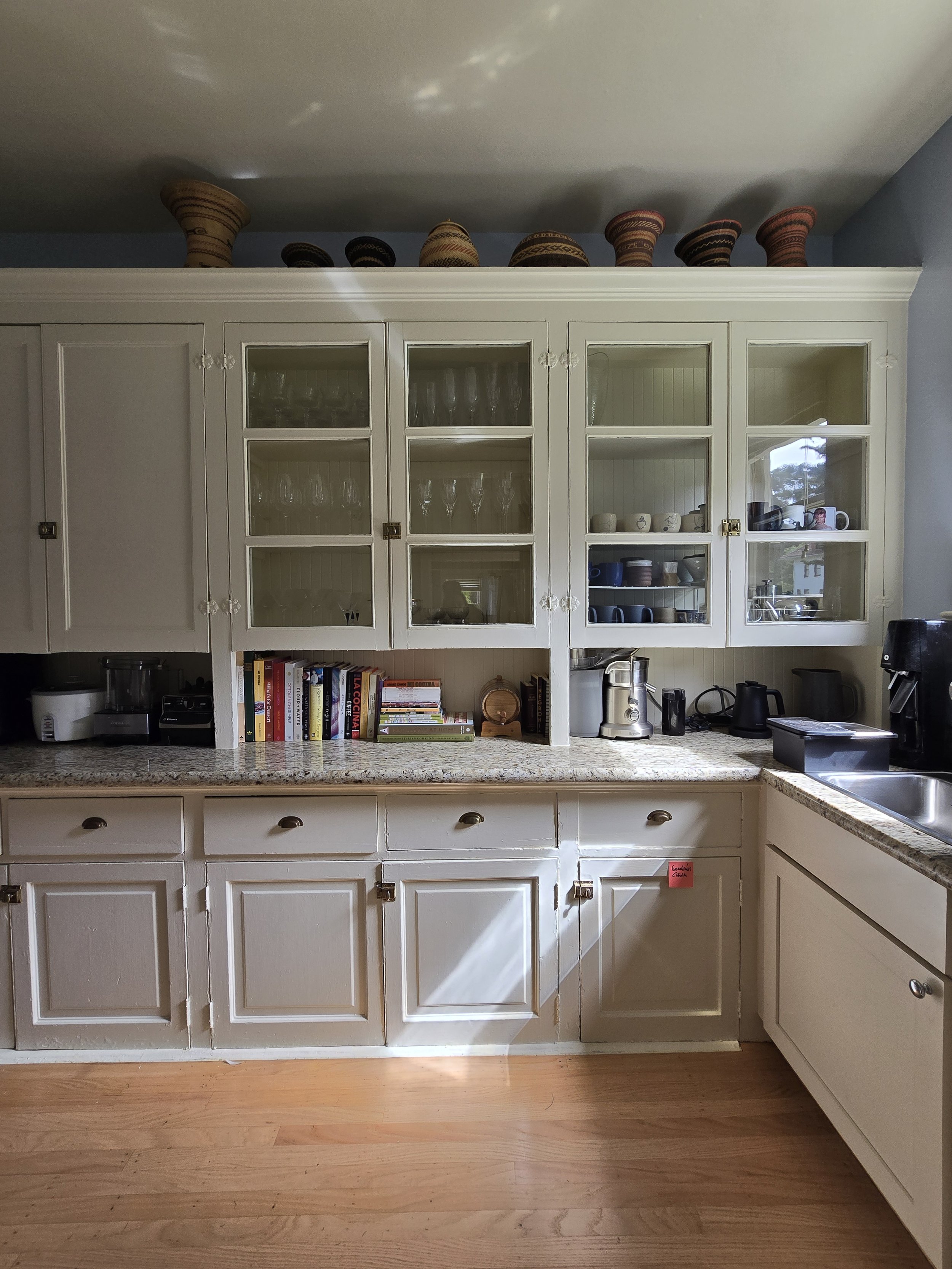 A kitchen with white cabinets, some with glass doors displaying glassware and dishes, a granite countertop, and shelves with books and small items. On the counter are small appliances, a timer, and a stainless steel pitcher. Above the cabinets are de