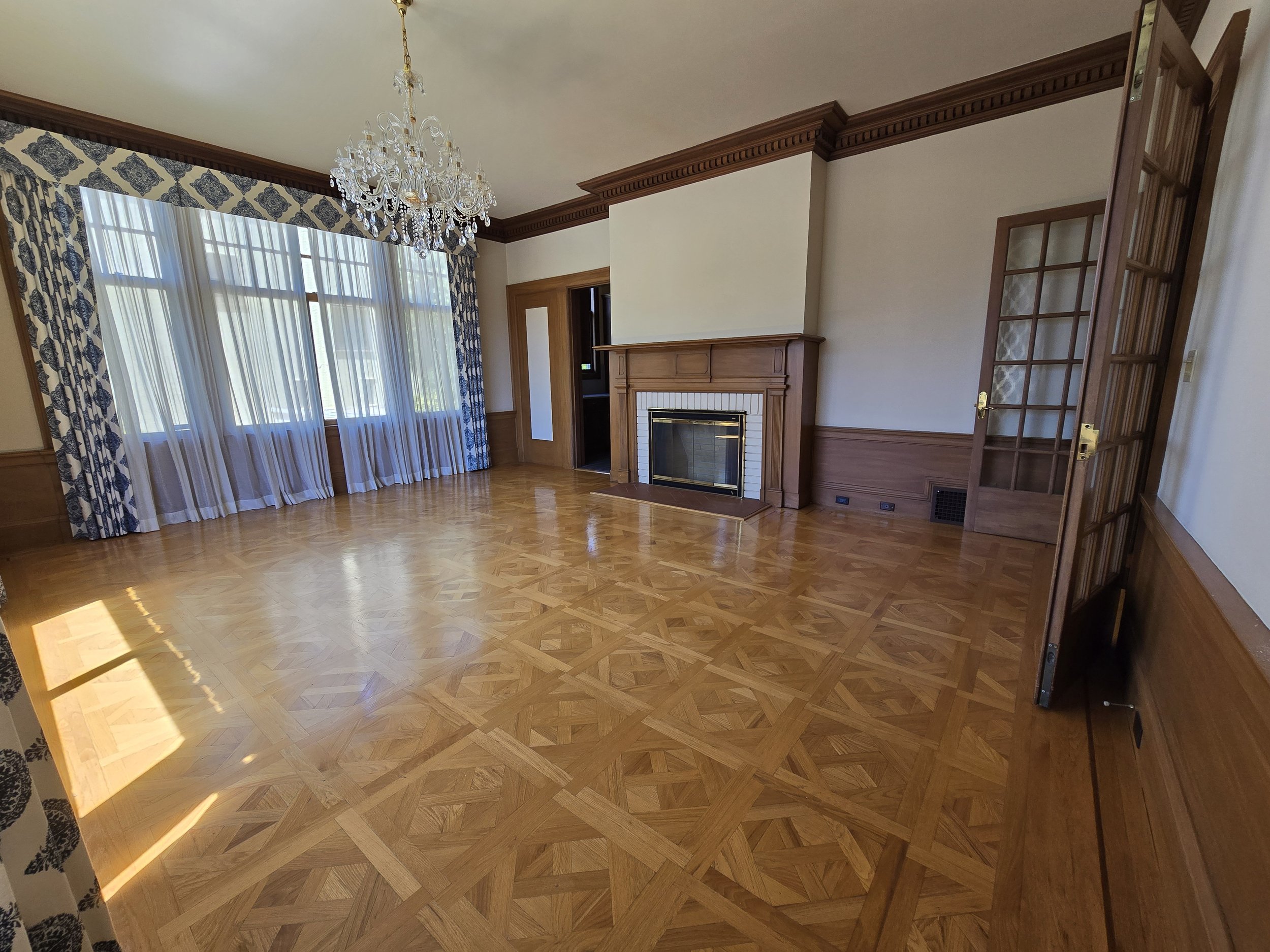 Empty living room with large window, patterned curtains, hardwood parquet floor, fireplace, chandelier, and wooden door with glass panels.