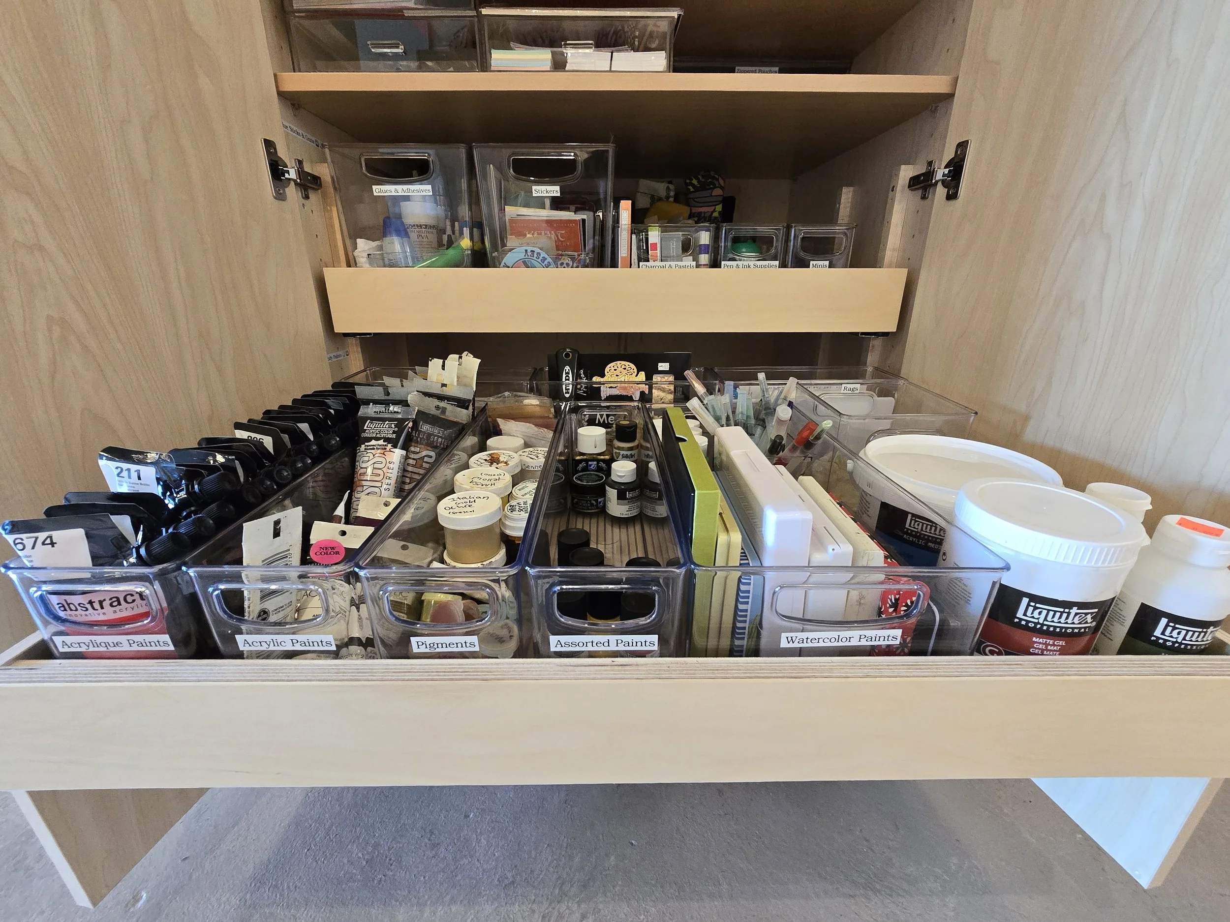 Open wooden art supply drawer containing bottles, tubes, and containers of acrylic paints, pigments, watercolor paints, and other art supplies organized in clear plastic containers.