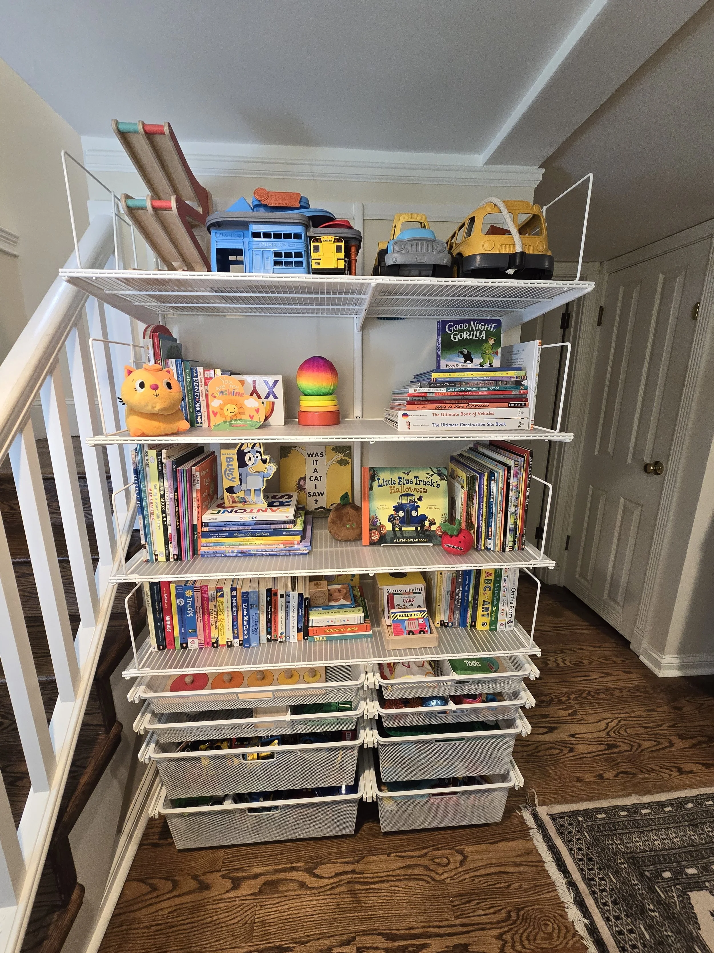 A white five-shelf storage unit filled with children's books, toys, and craft supplies, situated on a wooden floor next to a staircase and a beige wall with a closet door.