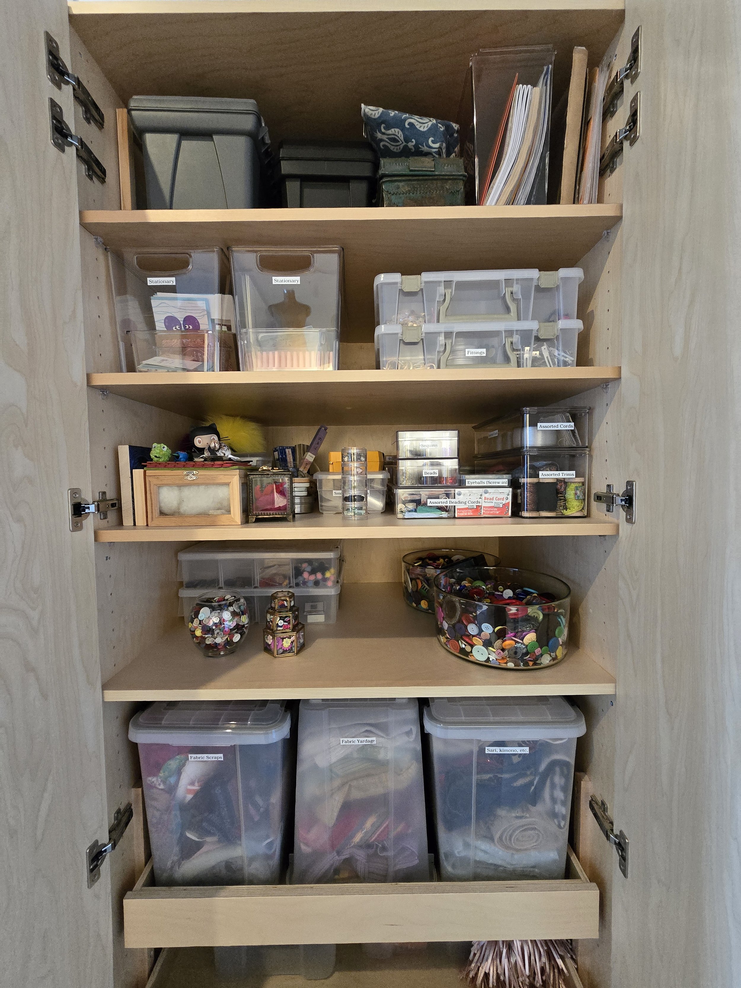 A wooden craft closet with labeled storage bins containing sewing and craft supplies, including fabric scraps, buttons, thread, and various small craft items.