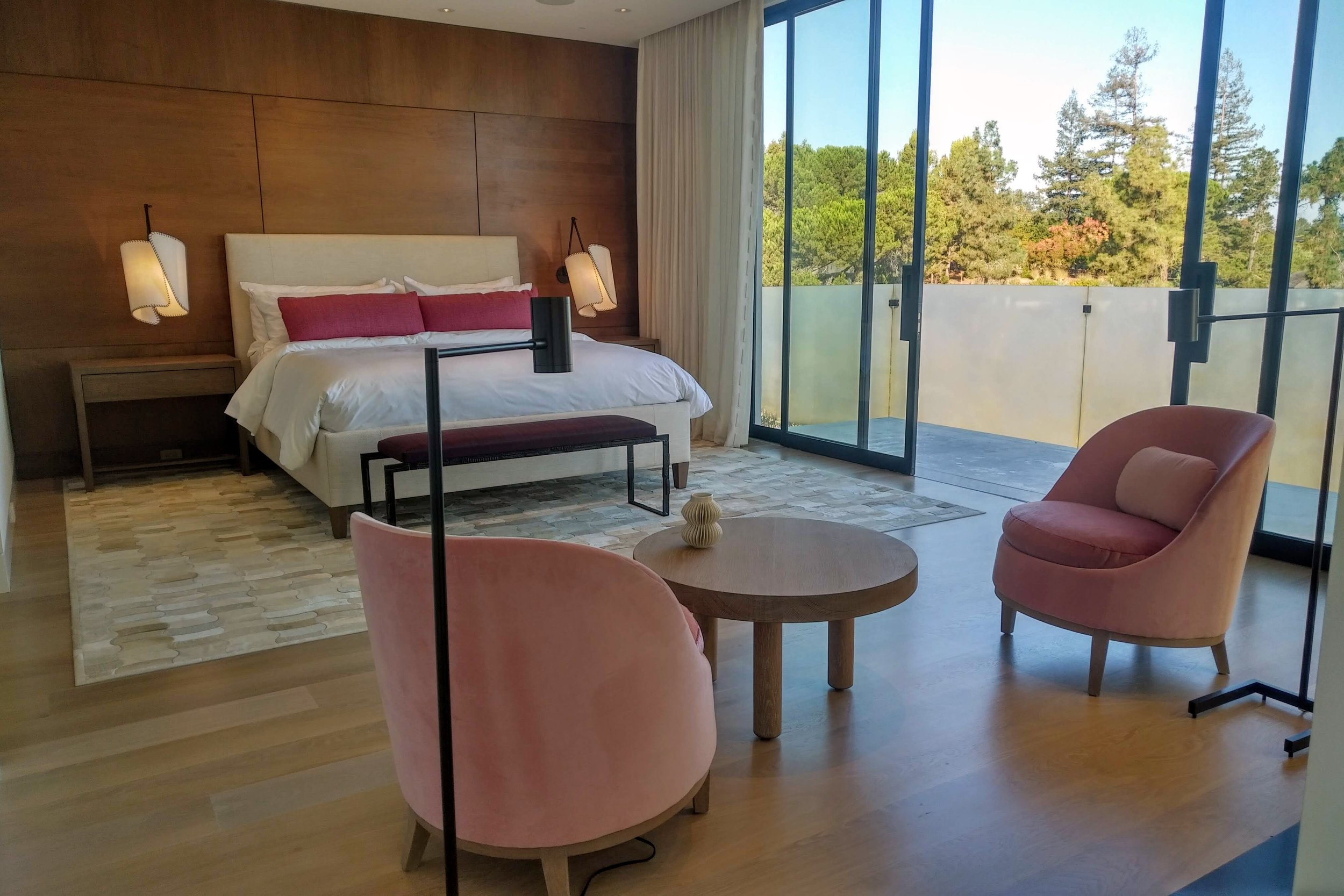 A modern bedroom with a large bed, pink pillows, bedside tables with hanging lamps, a pink armchair and a matching pink chair, a wooden coffee table, and a sliding glass door leading to a balcony with a view of trees.