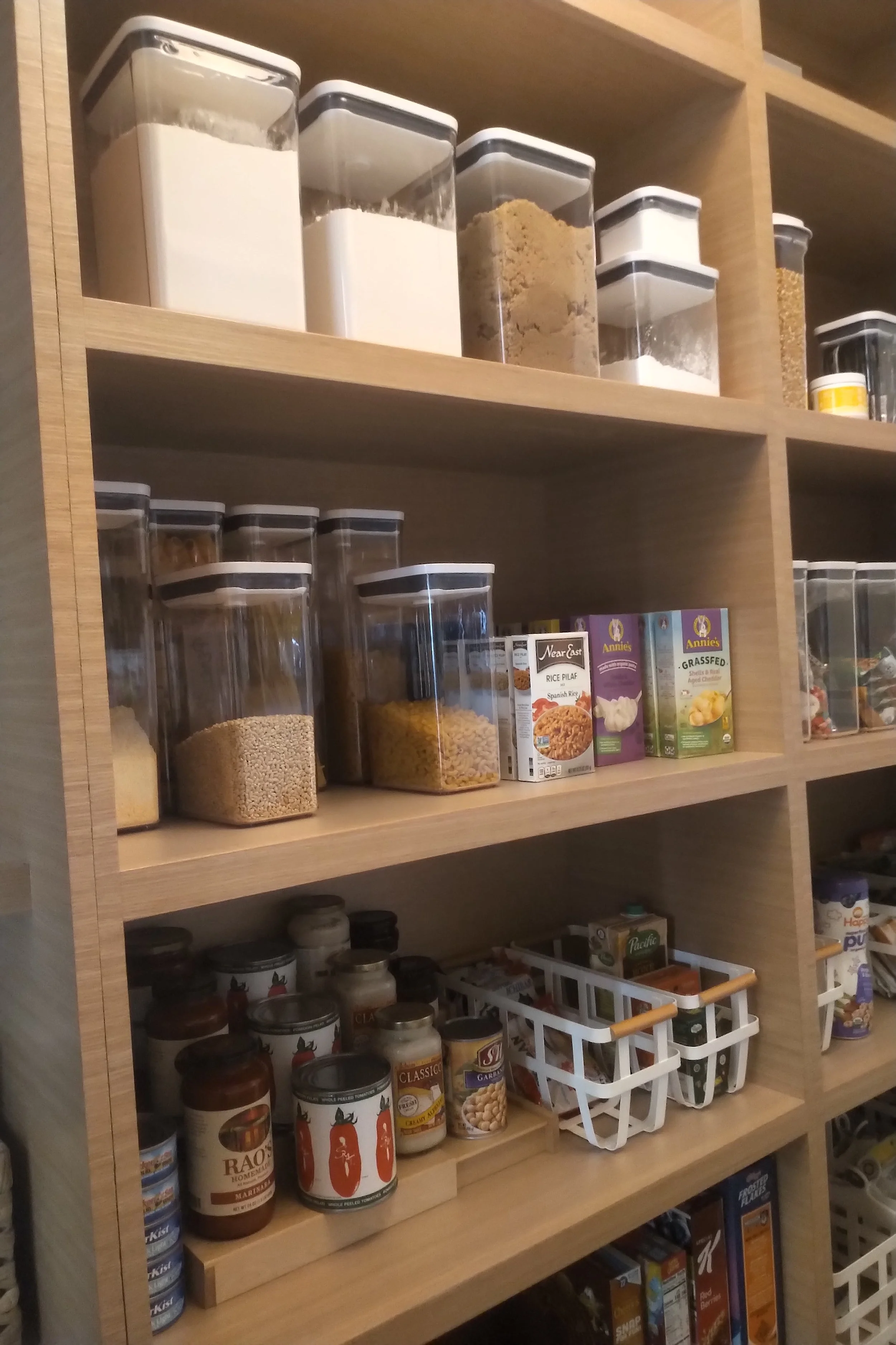 Organized pantry shelves with clear storage containers holding grains, powders, and snacks. Canned goods and boxed items are also visible.
