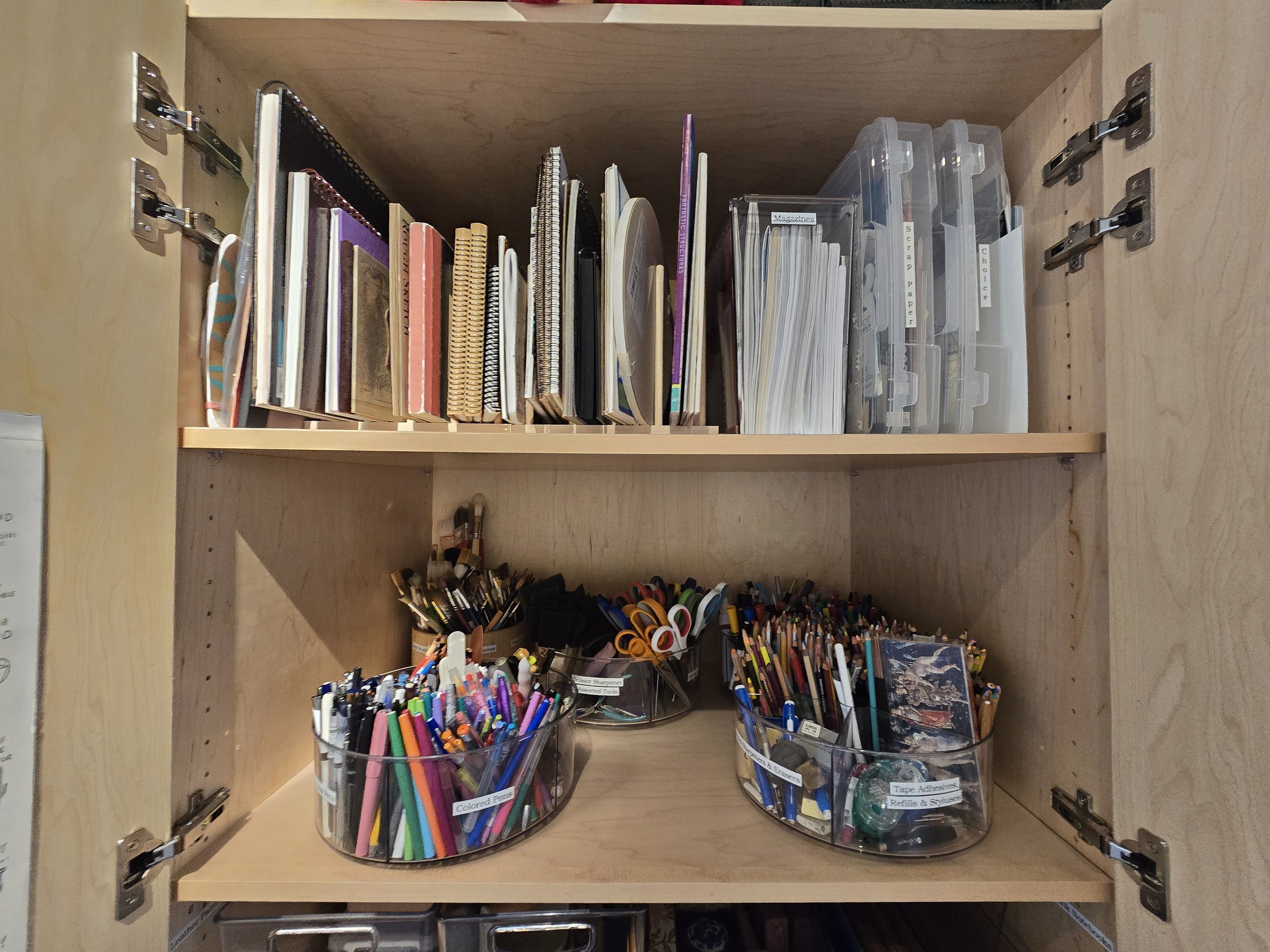 Open wooden desk drawer with two shelves. The top shelf contains various notebooks, binders, and paper organizers. The bottom shelf has two clear containers filled with colored pens, pencils, scissors, and other stationery supplies.