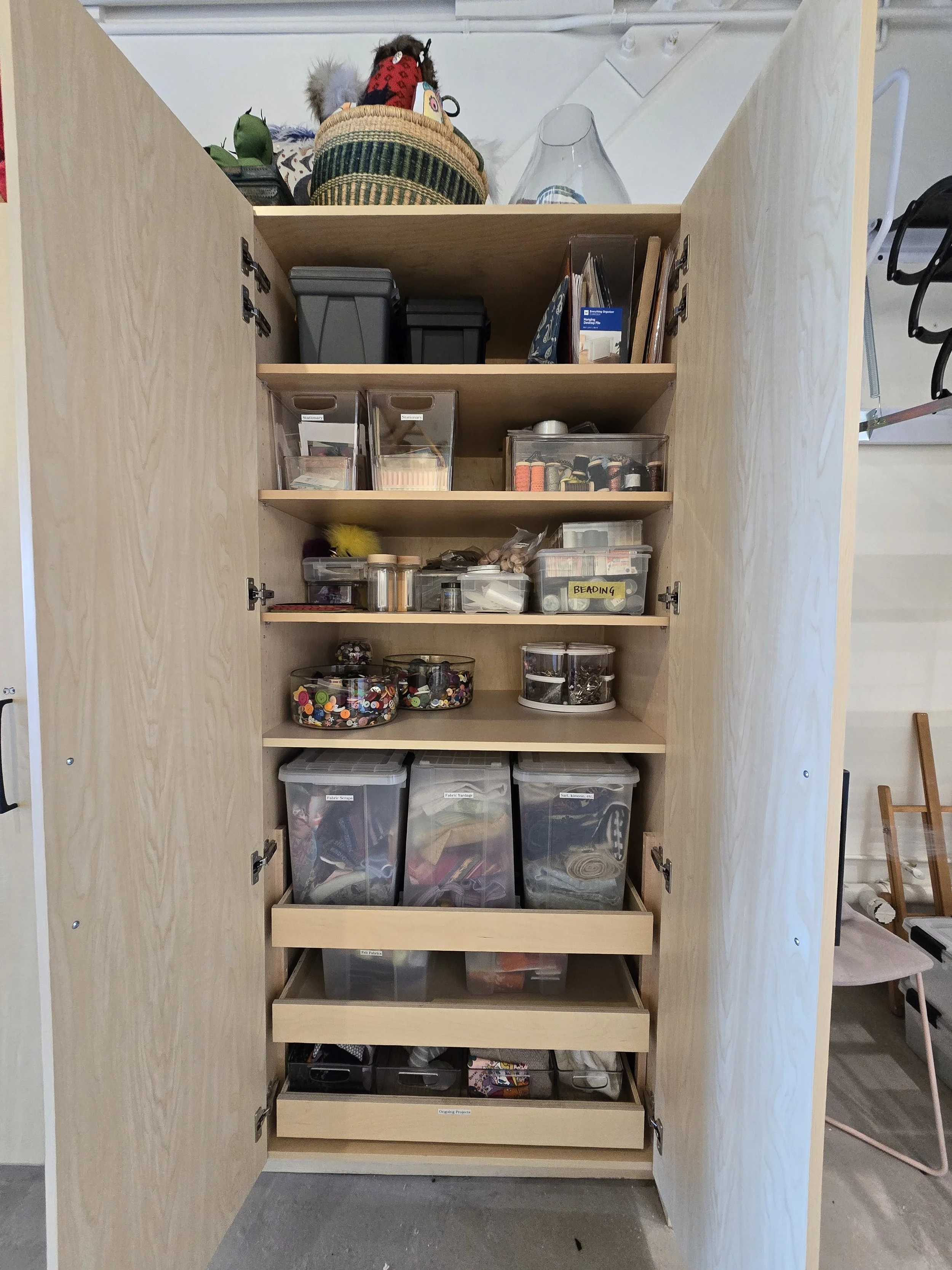 A wooden storage cabinet with open doors, containing organized craft and sewing supplies, including clear bins with buttons, thread spools, ribbons, and fabric.
