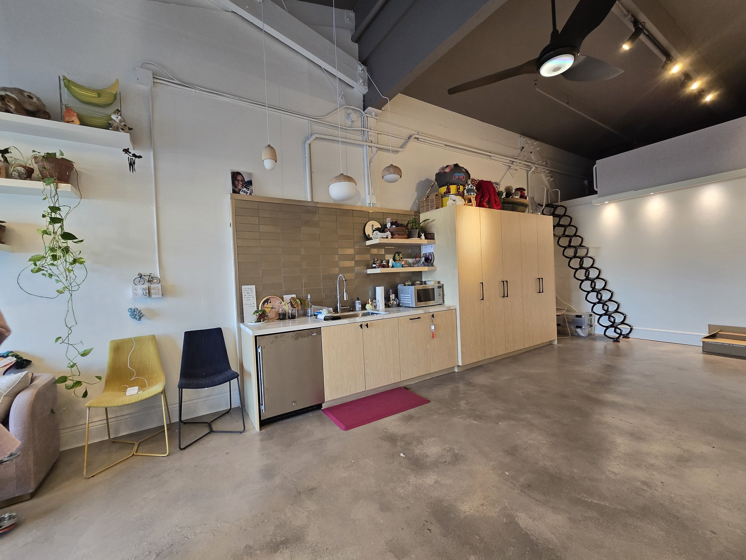 Open kitchen with wooden cabinets, microwave, mini fridge, and a sink. Three chairs are positioned against the wall, two are yellow and one is black. There are various items on the shelves and counter, and a staircase with a black spiral design leadi