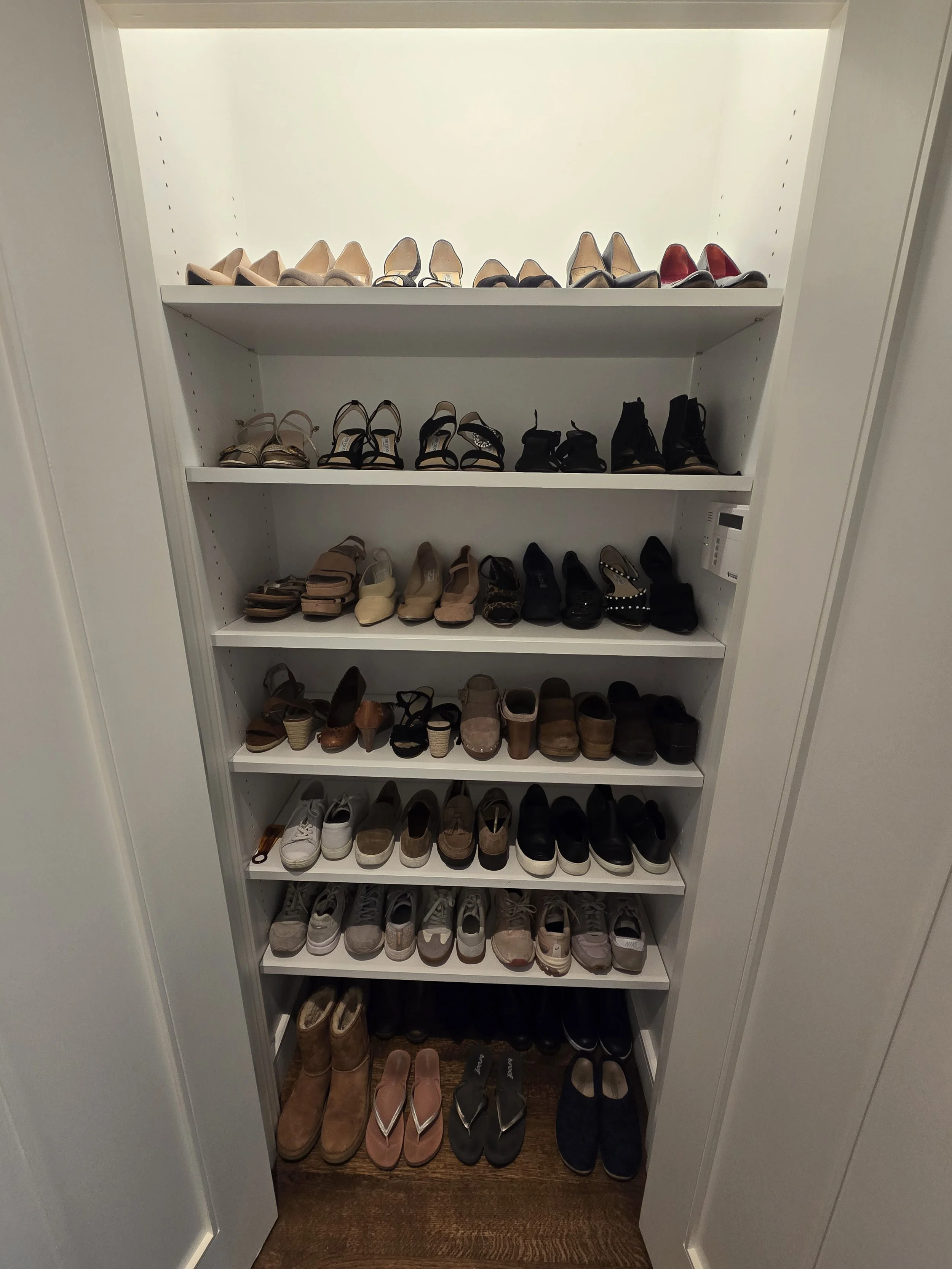 A white shoe cabinet with multiple shelves filled with various women's shoes, including high heels, sandals, sneakers, boots, and flats.