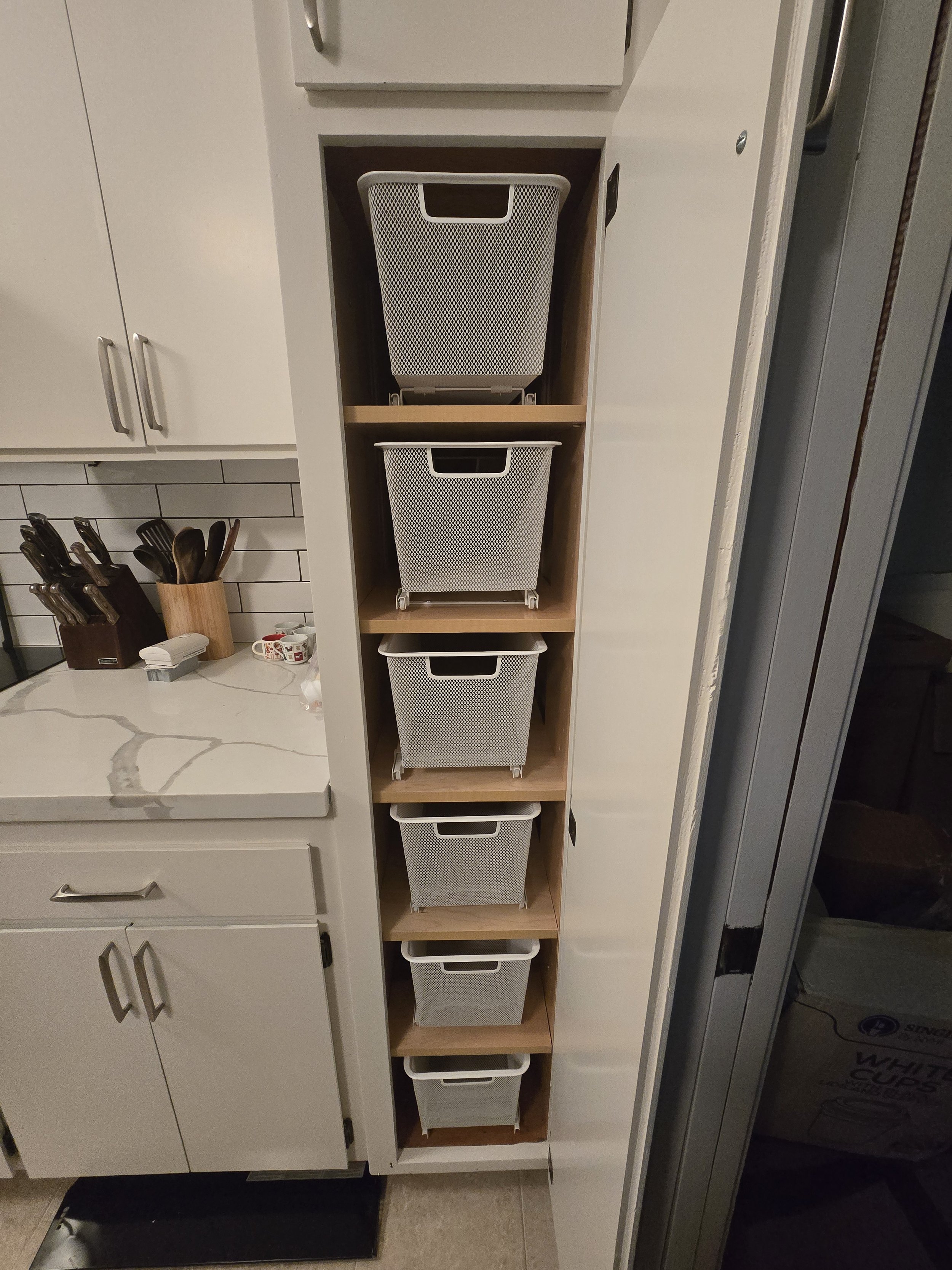 A narrow bookshelf with five shelves holding white mesh storage baskets in a kitchen.