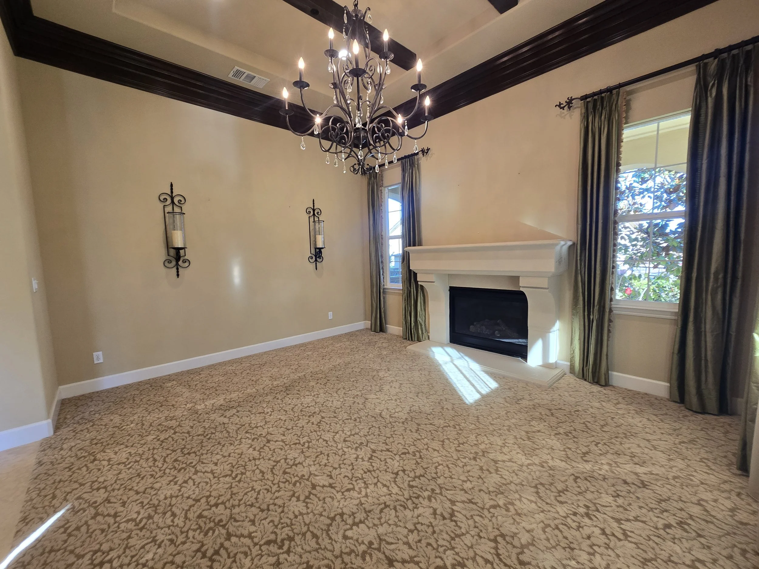 Empty living room with beige walls, a chandelier, wall-mounted candle holders, a fireplace, and windows with green curtains.