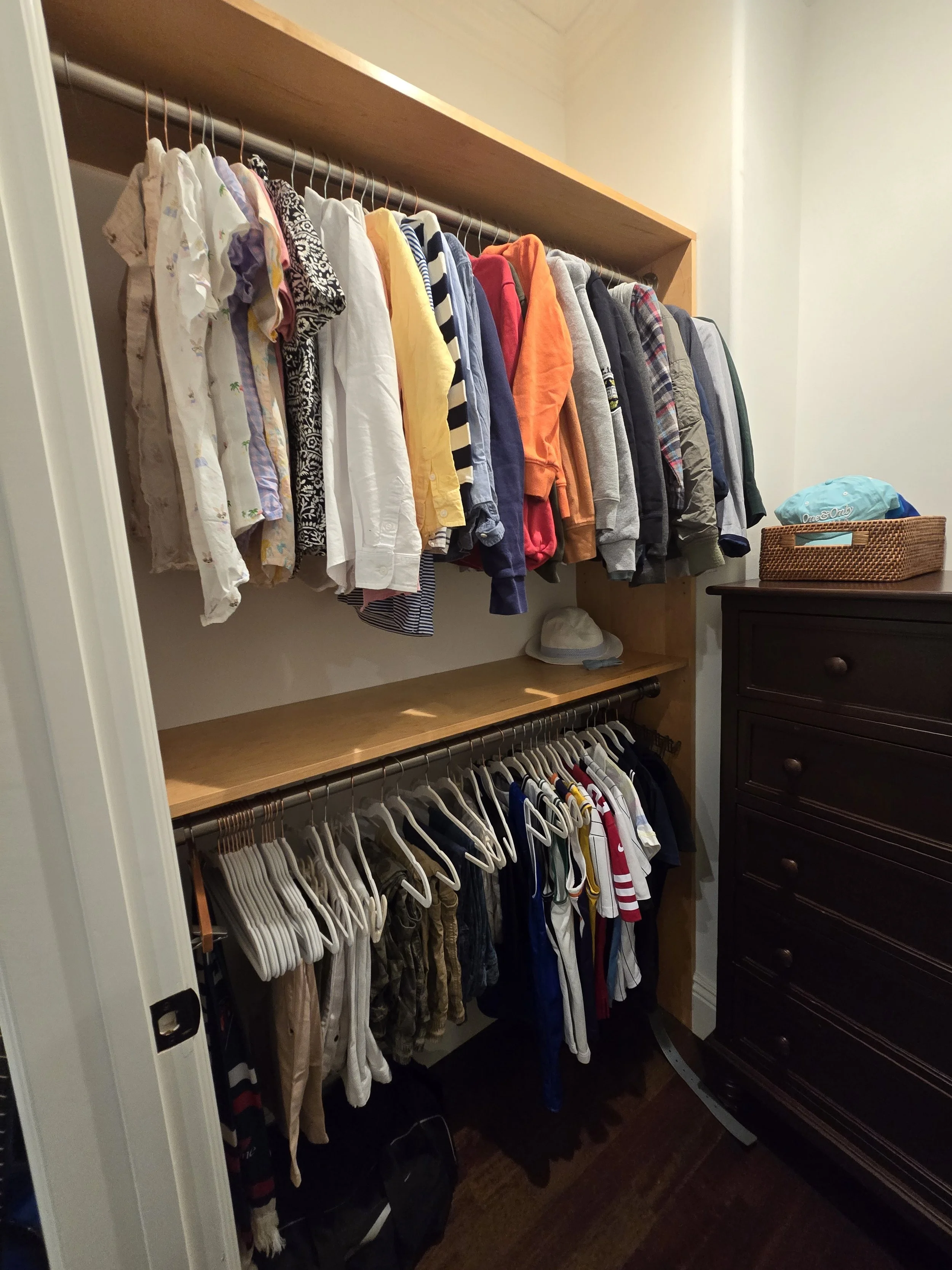 A closet with two shelves of hanging clothes, a hat on the middle shelf, and a dresser on the right with a basket on top.