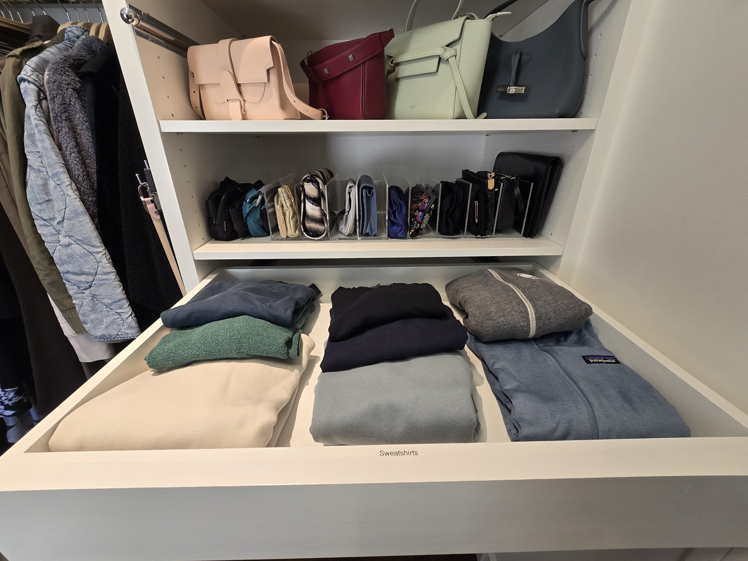 Display of folded sweatshirts on a white table, with a label reading 'Sweatshirts', and a shelf above holding handbags and purses of various colors and sizes.