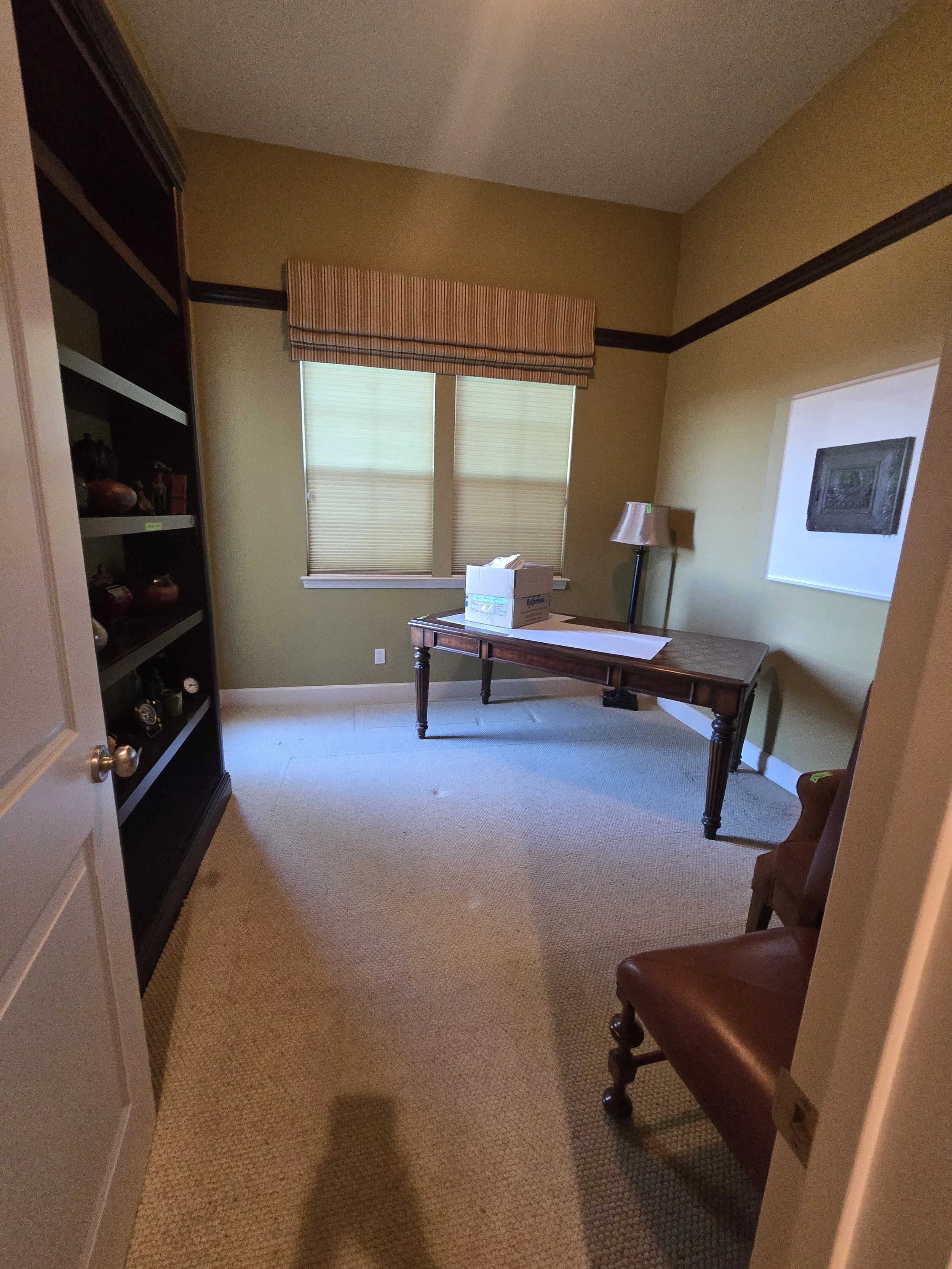 Empty room with a wooden desk, a box, a floor lamp, a shelf with decorative items, and a window with blinds and a valance.