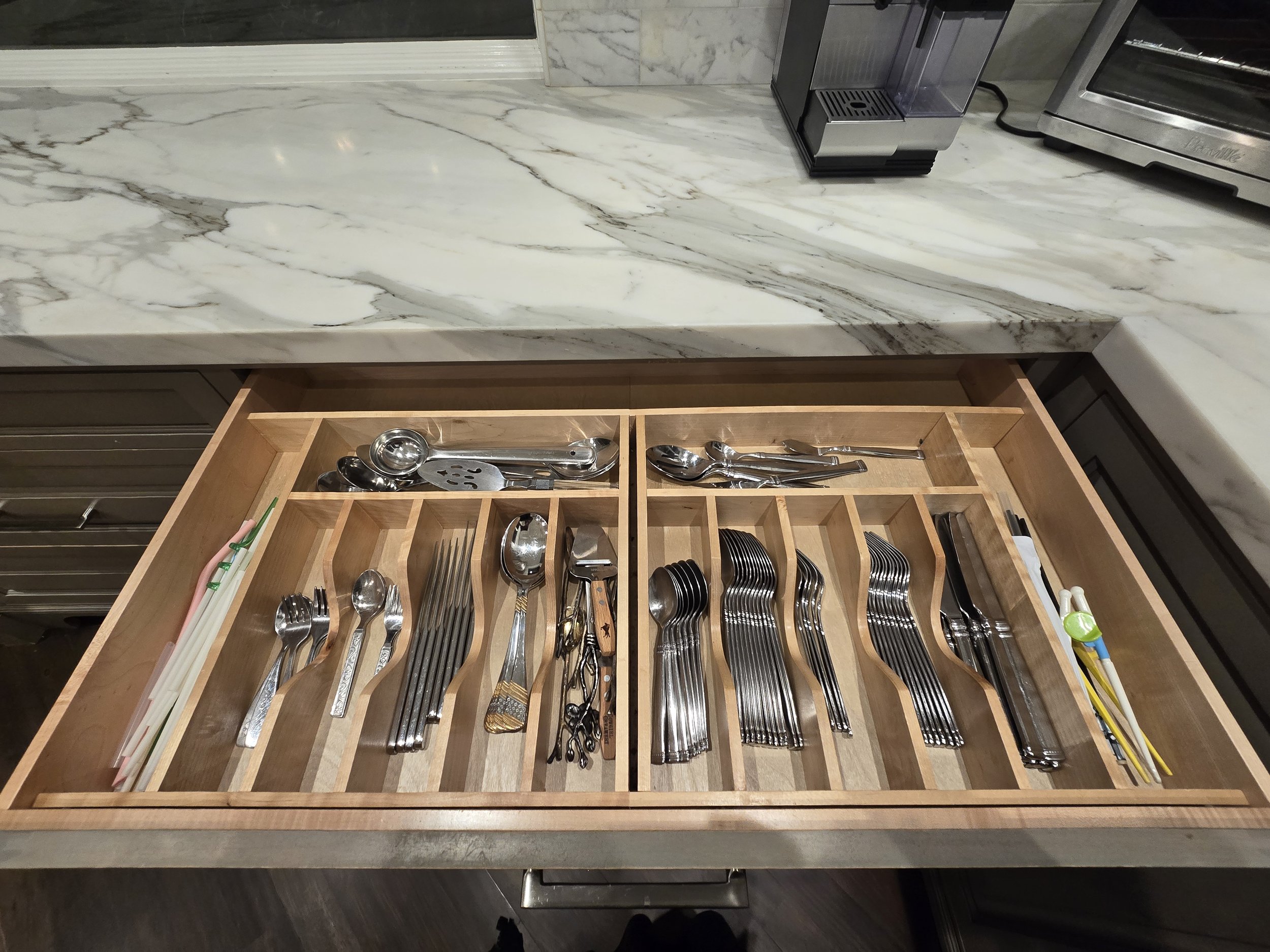 Open kitchen drawer with organized utensils including forks, spoons, knives, chopsticks, and small kitchen tools on a marble countertop.