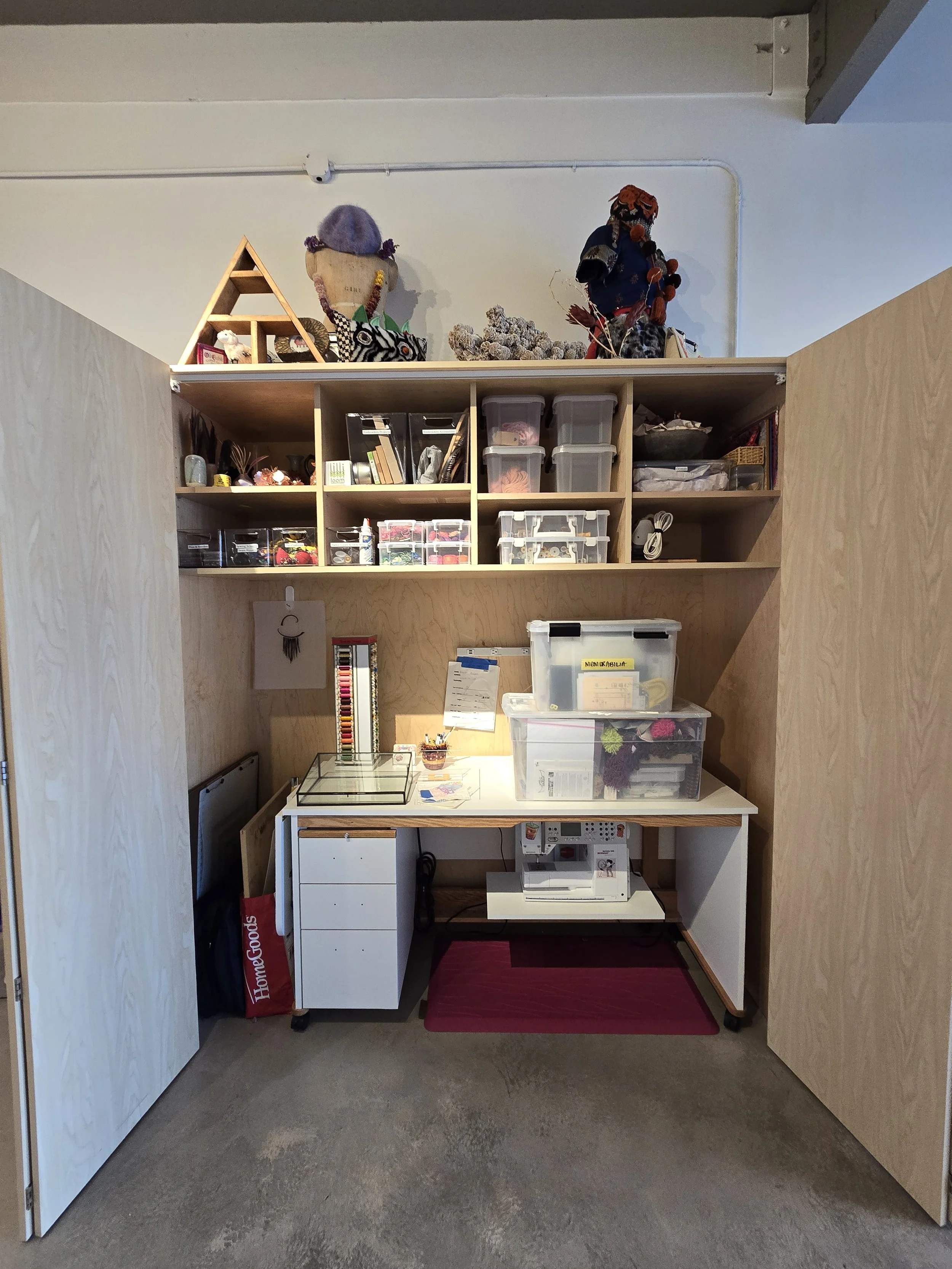 A workspace with a wooden shelving unit filled with craft supplies, containers, and figures. A small table with drawers, a sewing machine, and a red mat are in front of the shelves. The room has plain beige walls and a concrete floor.
