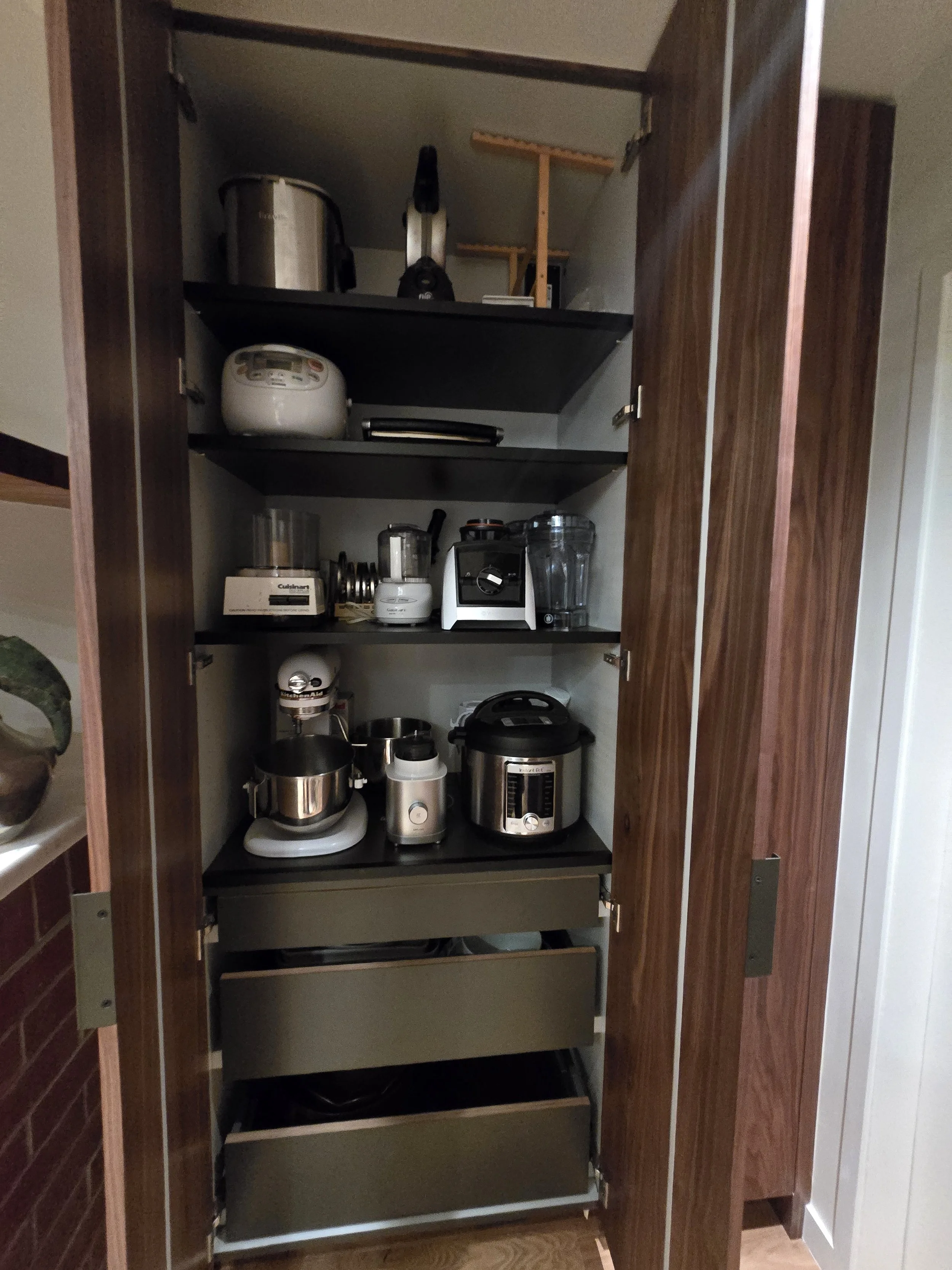 An open kitchen cabinet with various appliances and kitchenware, including a rice cooker, blender, and toaster, stored on shelves and inside drawers.