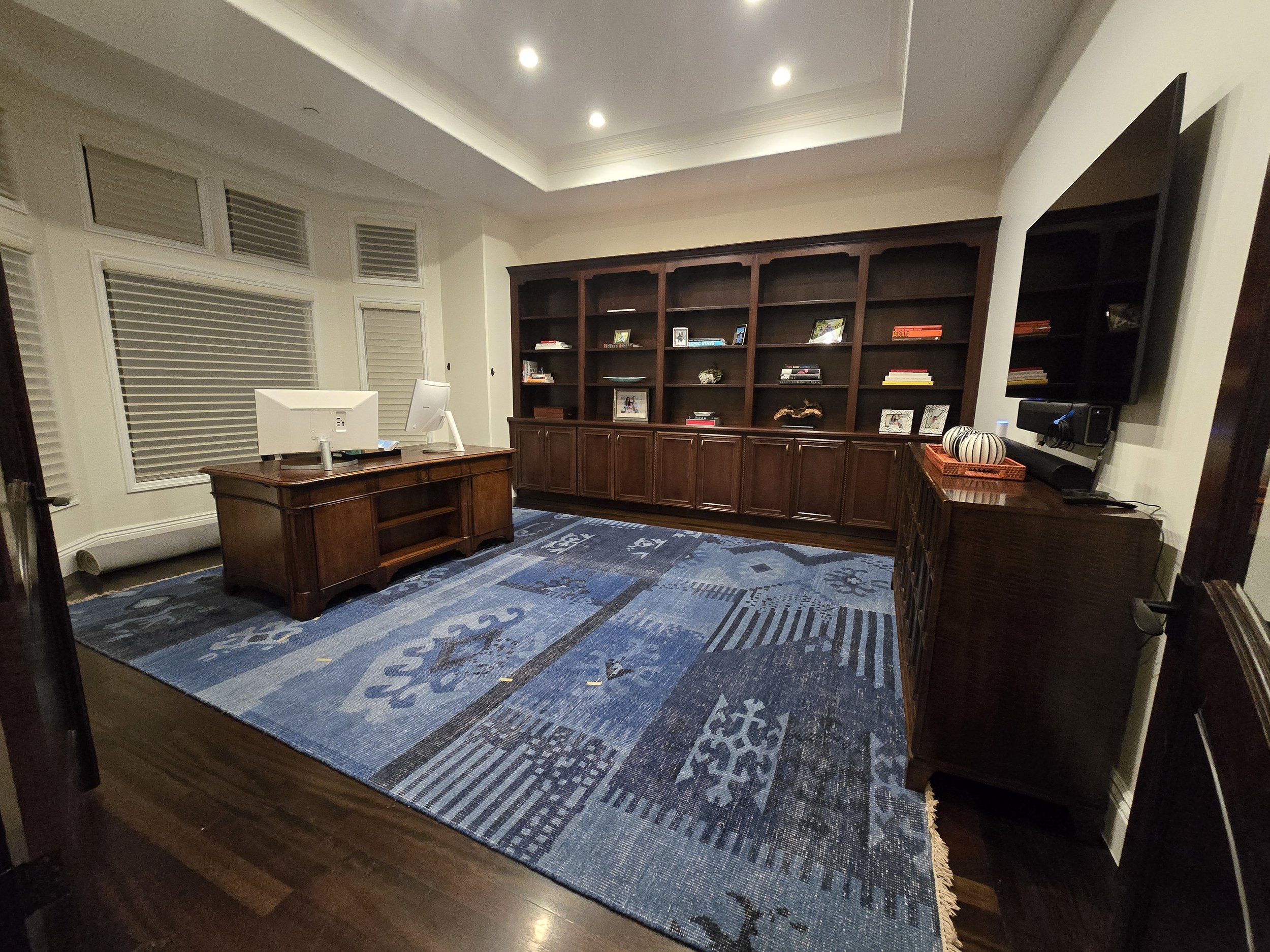 An office room with a wooden desk, white computer monitors, a large wooden bookshelf, a mounted flat-screen TV, and a patterned blue rug on hardwood flooring.