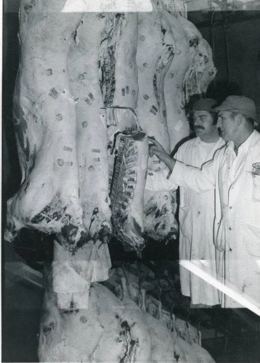Black and White photograph of 2 butchers looking at hanging beef