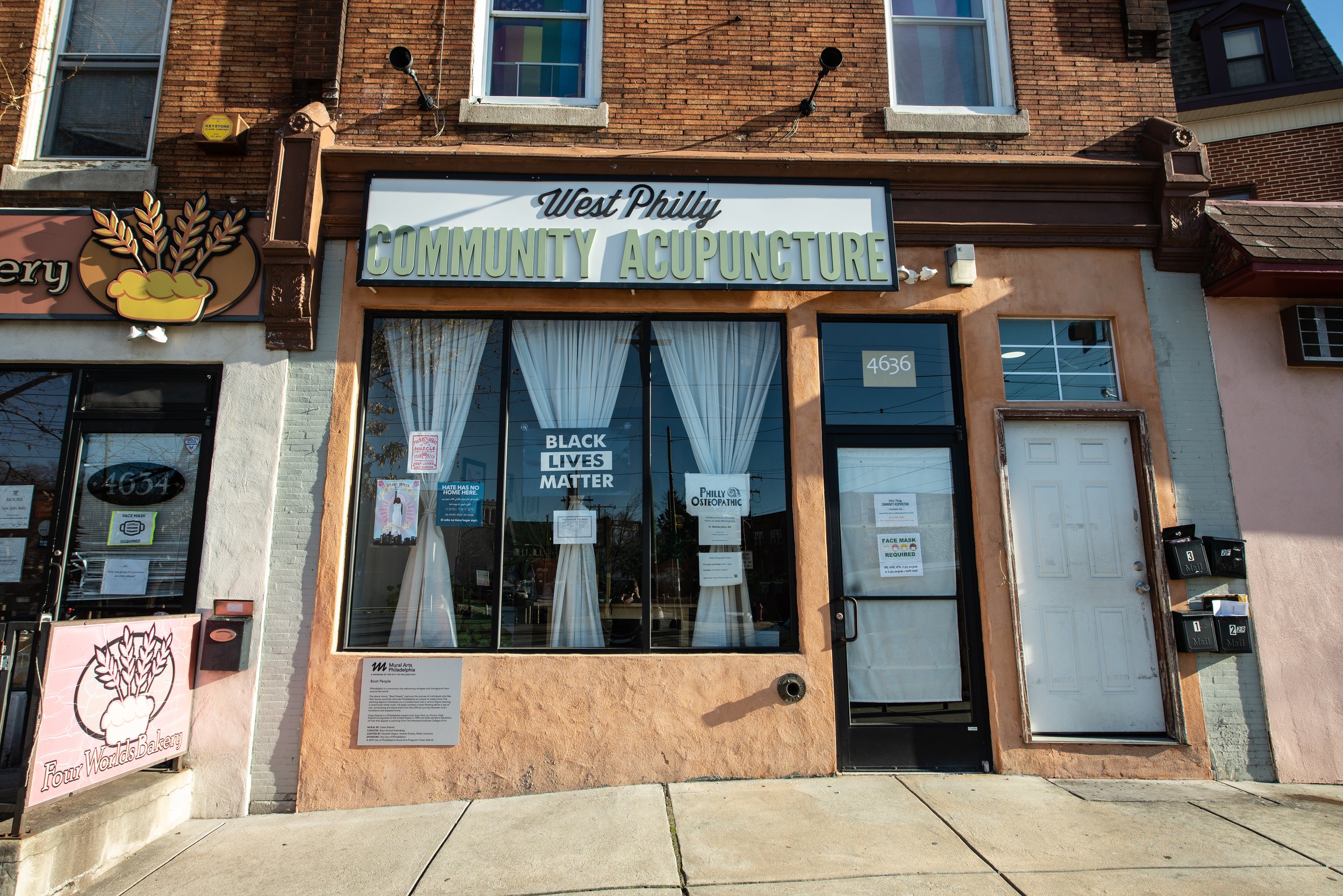  image description: The exterior of a brick building. A sign is hung over three large windows and the front door that reads ‘West Philly Community Acupuncture’ in a bold font.  