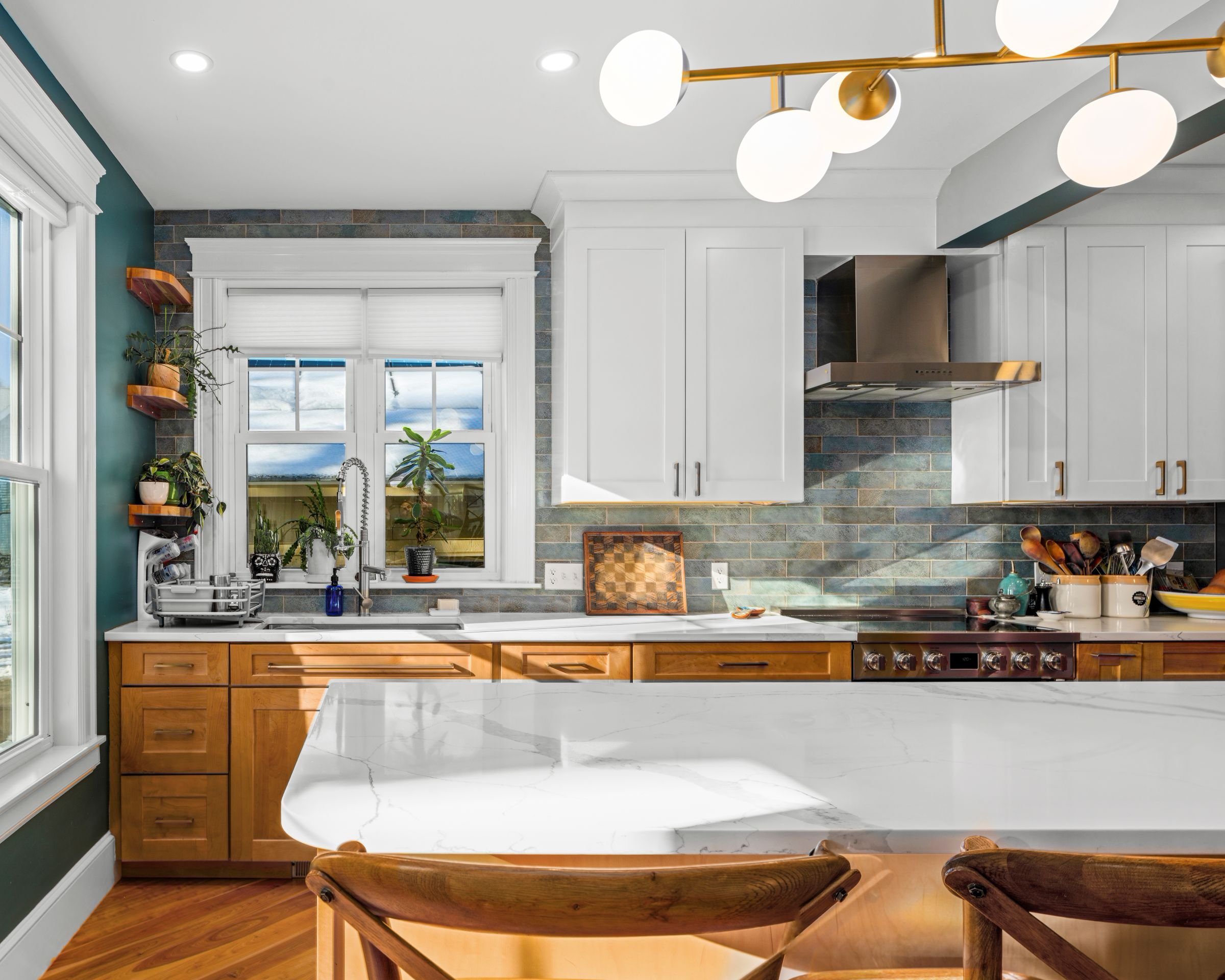 Wide-angle view of a Worcester MA kitchen remodel featuring white upper cabinets, natural wood lowers, Statuario Classic quartz countertops, Kalay Green tile backsplash, and a central island with seating.