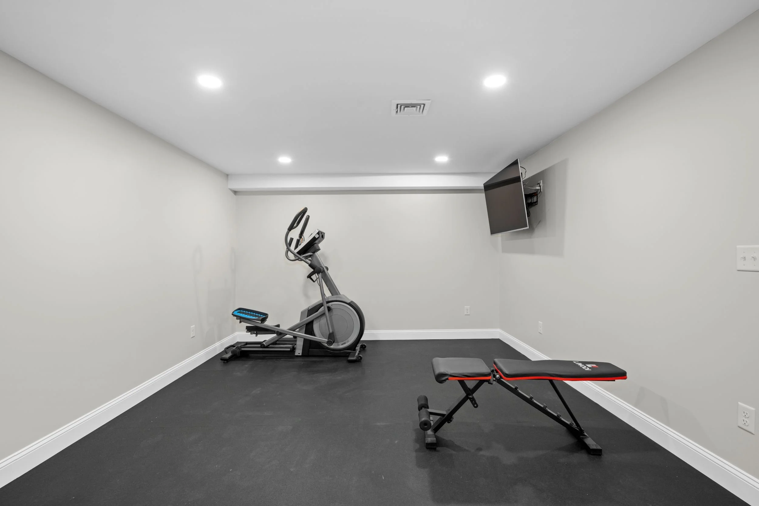 Dedicated home gym in a finished basement with professional rubber flooring, wall-mounted mirrors, and recessed LED lighting in Boxborough, MA.