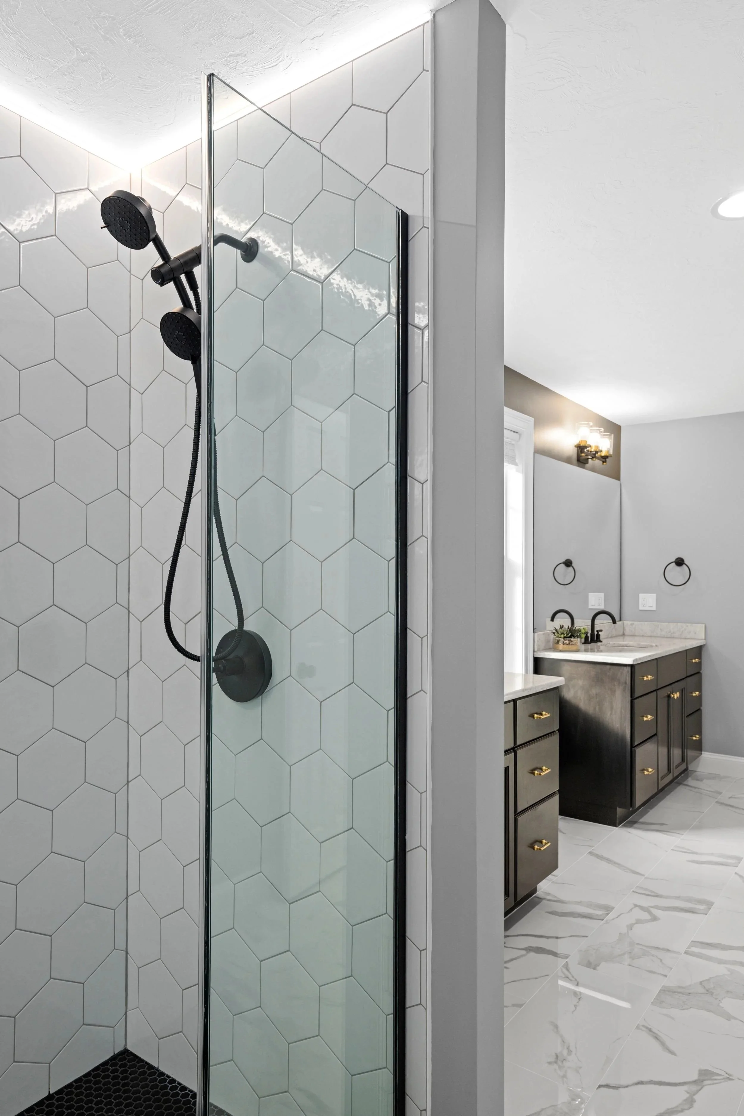 Frameless glass shower panel with white hex tile and matte black fixtures, viewed toward the double vanity in a Boxborough MA master bathroom remodel
