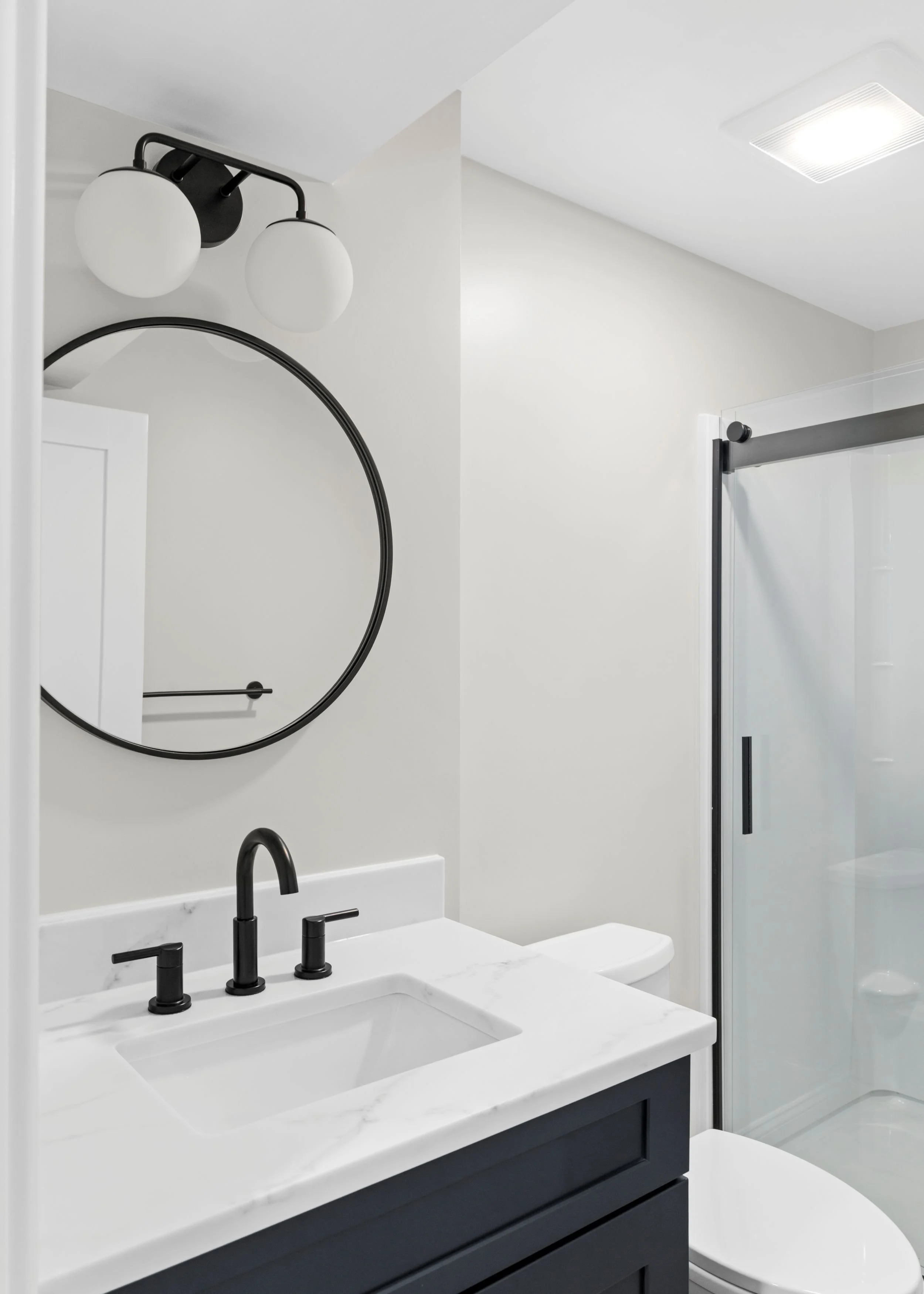Modern basement bathroom in Boxborough, MA featuring a charcoal grey vanity, white quartz countertop, and a matte black round mirror.