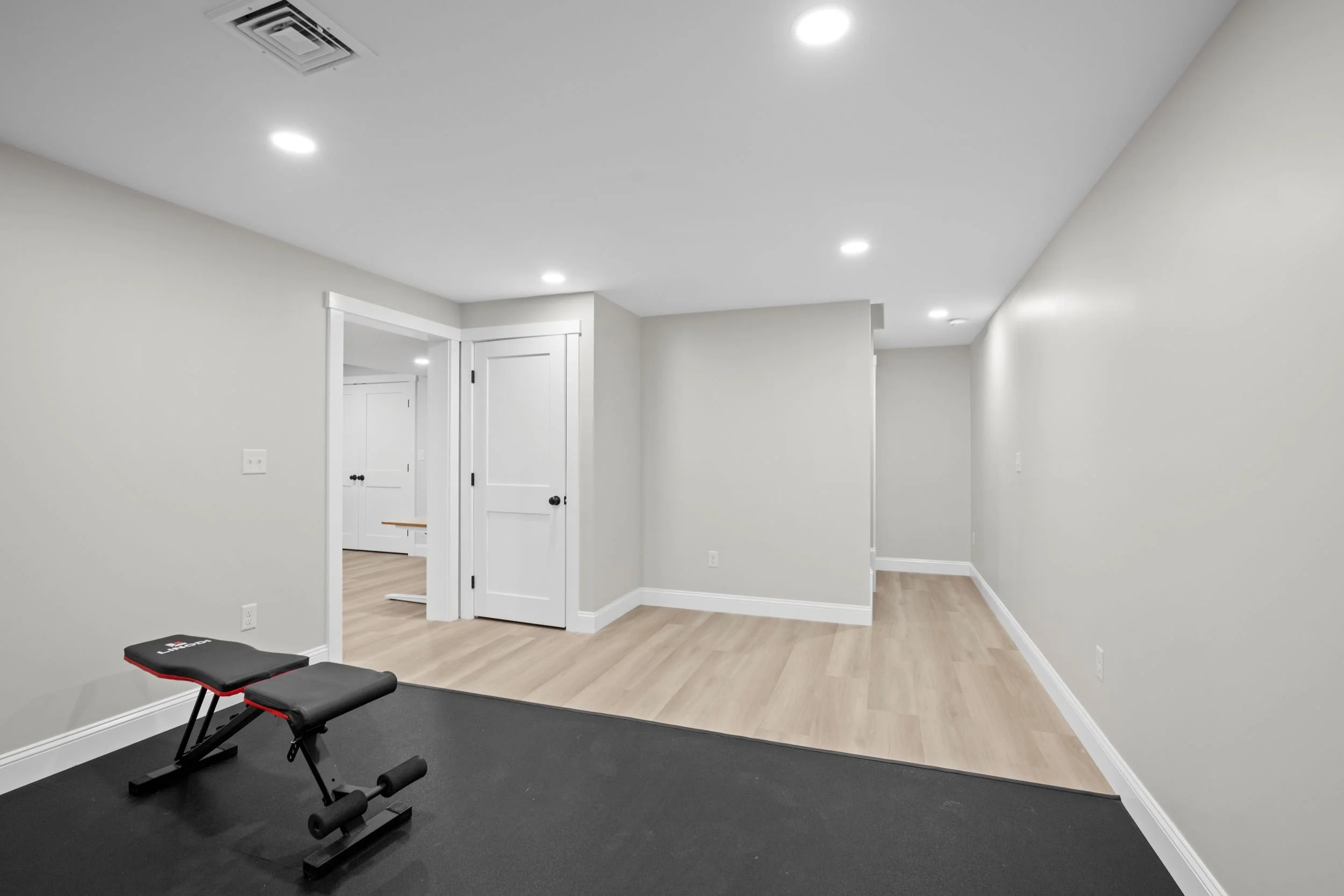 right, finished basement bonus room in Boxborough, MA featuring light grey walls, white shaker doors, and a transition from rubber gym flooring to luxury vinyl plank.