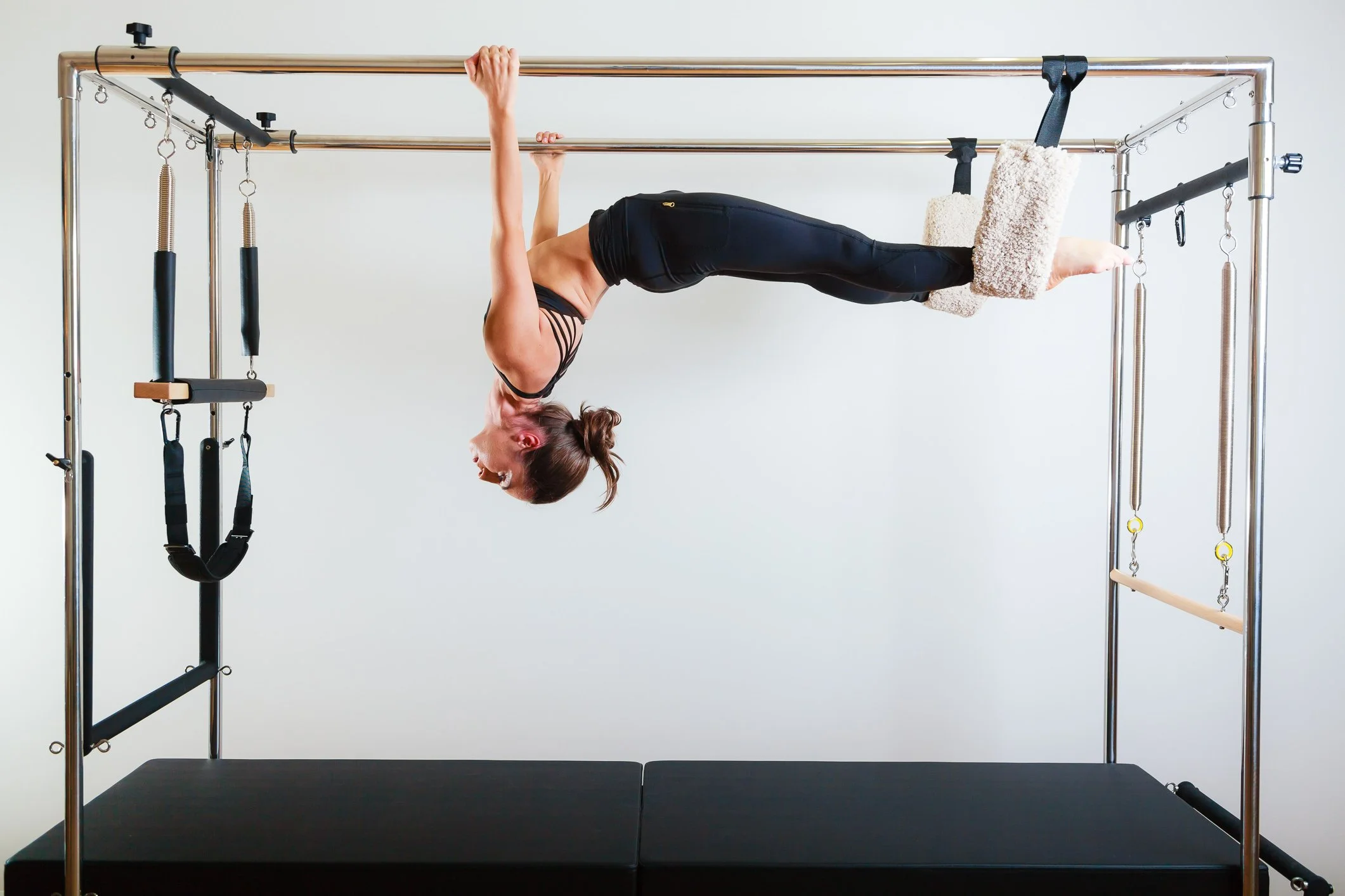A client performing Hanging Back on the Cadillac, suspended upside down by the ankle straps and gripping the trapeze bar.