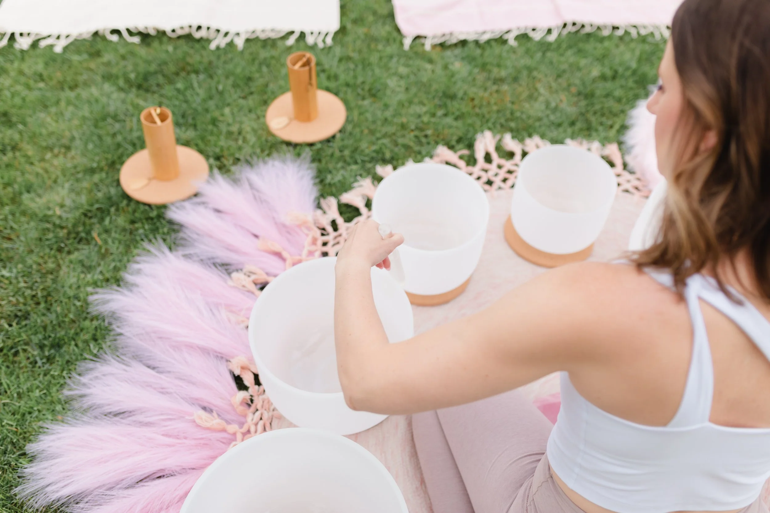 Woman seated on grass playing crystal singing bowls during sound therapy session.