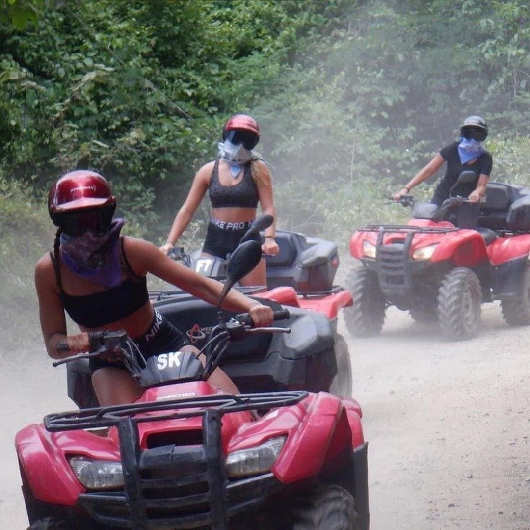 Three women on ATVs with bandanas riding dusty jungle trail.