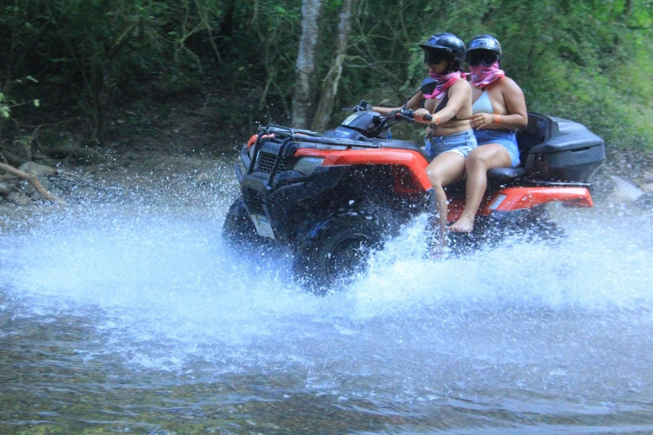 Two guests riding double on ATV crossing river with My Deztination.