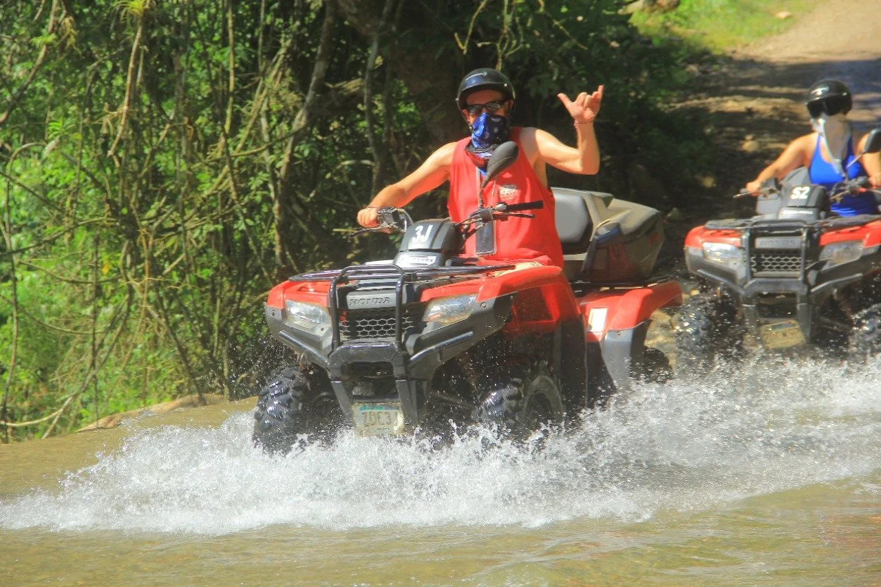 Guest raising hand riding ATV through water with My Deztination.