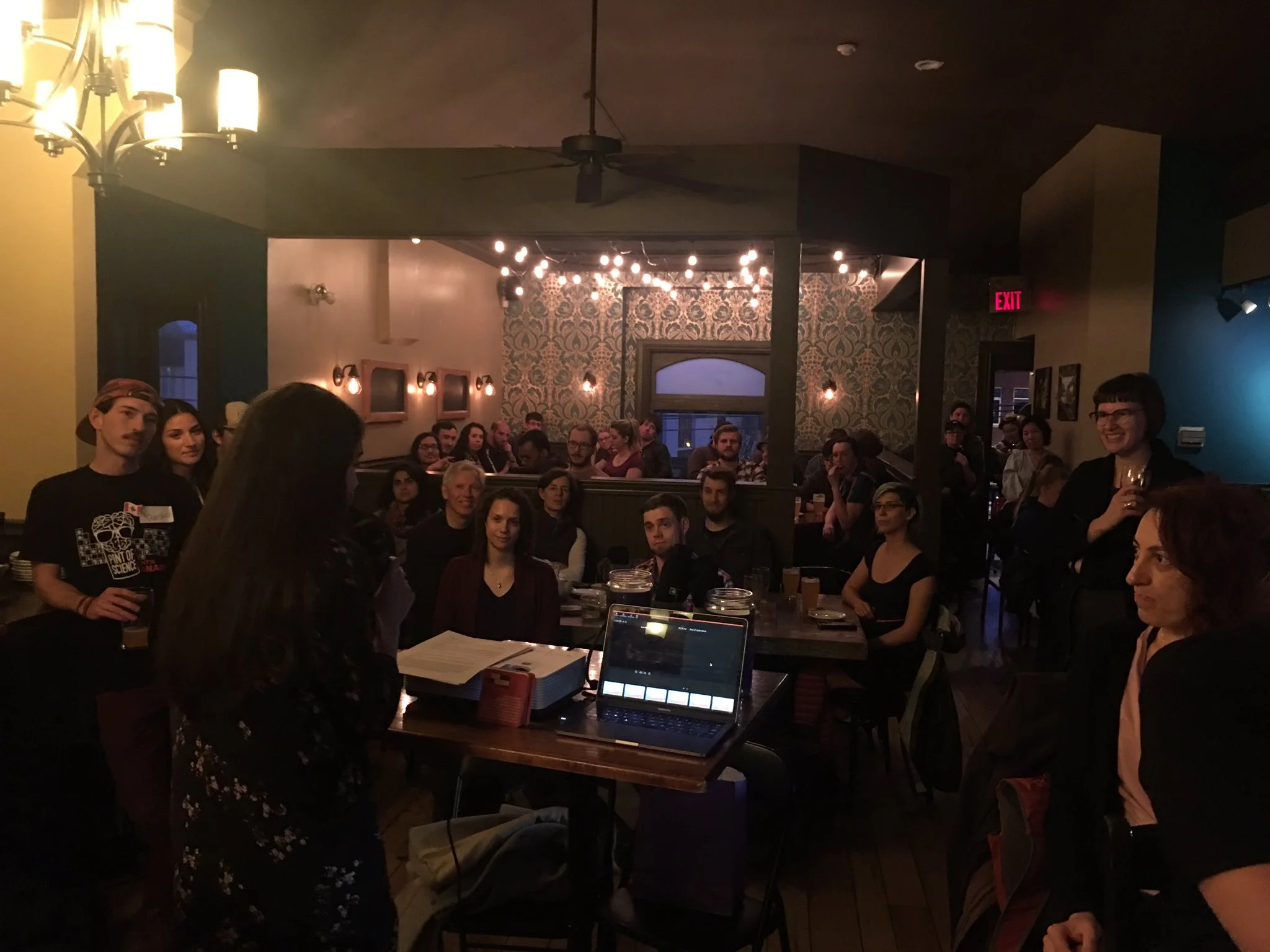 I am standing in a pub filled with people, with my dark hair down, wearing a black top with pink flowers and black jeans.