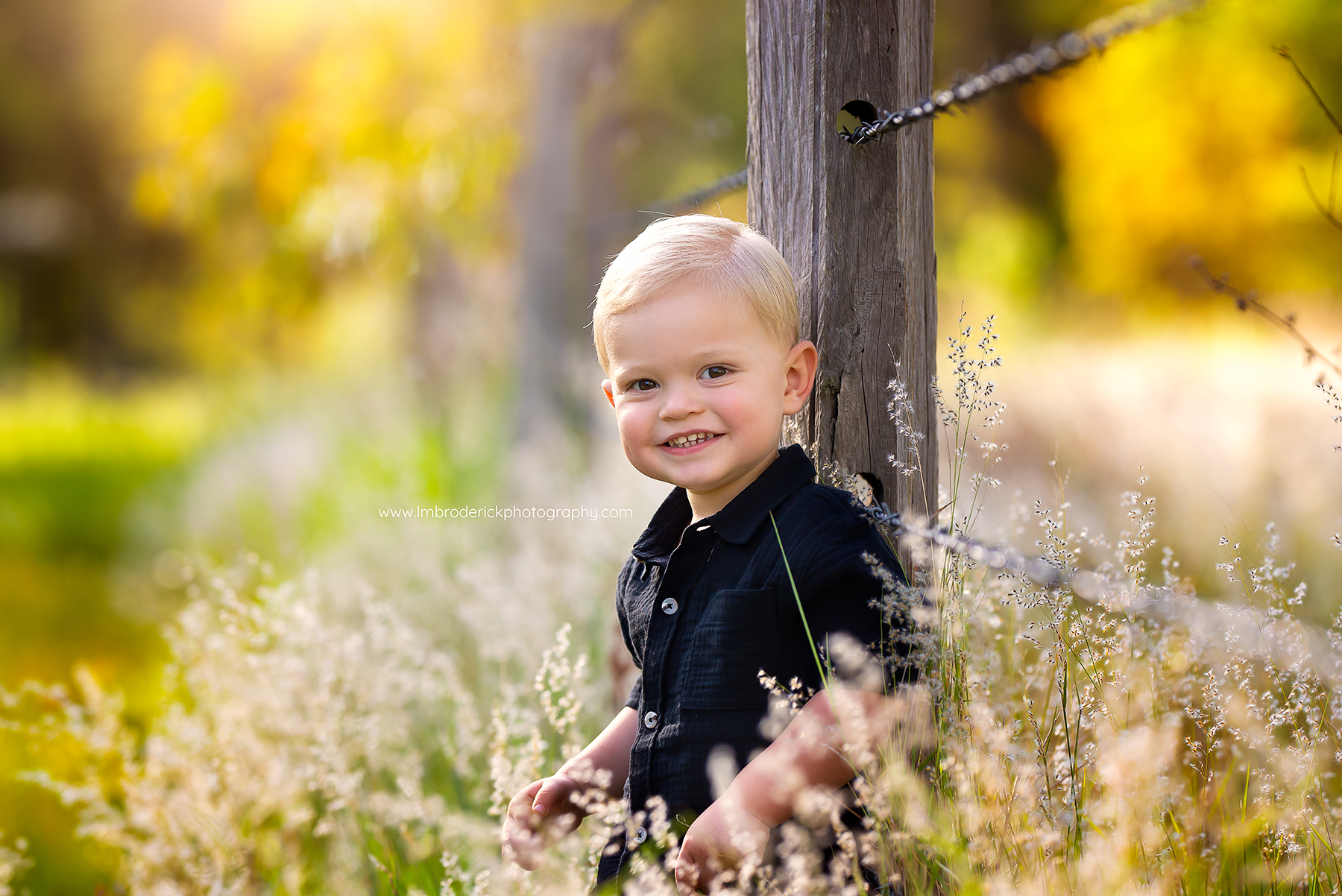 Young Fernvale boy sitting against a fence in long grass smiling
