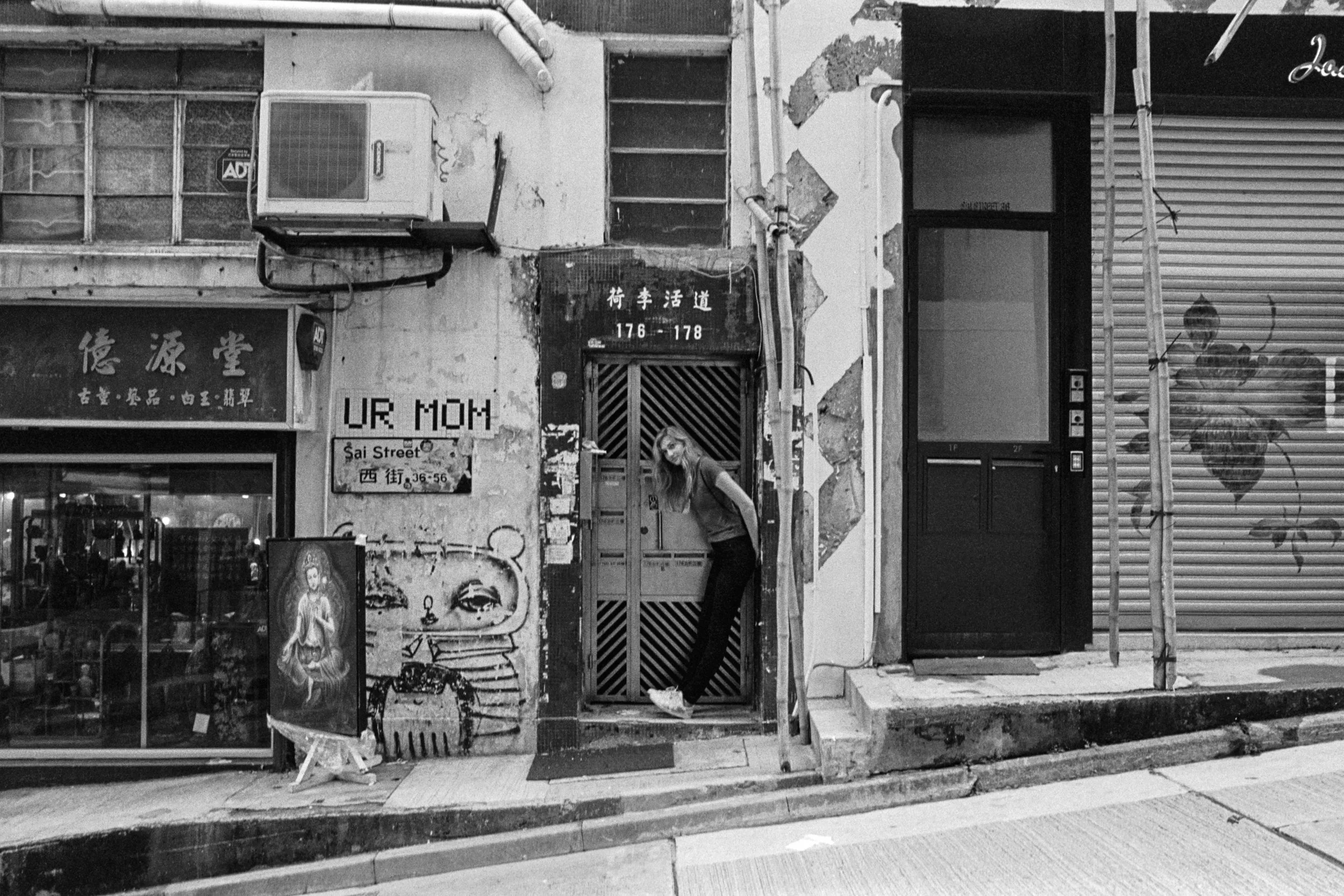Kodak Tri-X 400 @ 28mm Focal Length - Interacting with the environment by leaning against a doorway. Compositional variables include rectilinear lines of doorways, buildings, and an incline on the road.