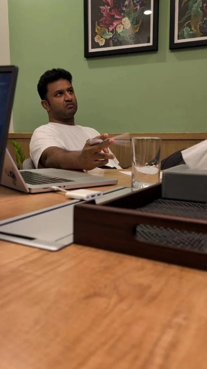 A man sitting at a conference table in a room with green walls, holding a smartphone, with a laptop, a glass of water, and office supplies on the table.