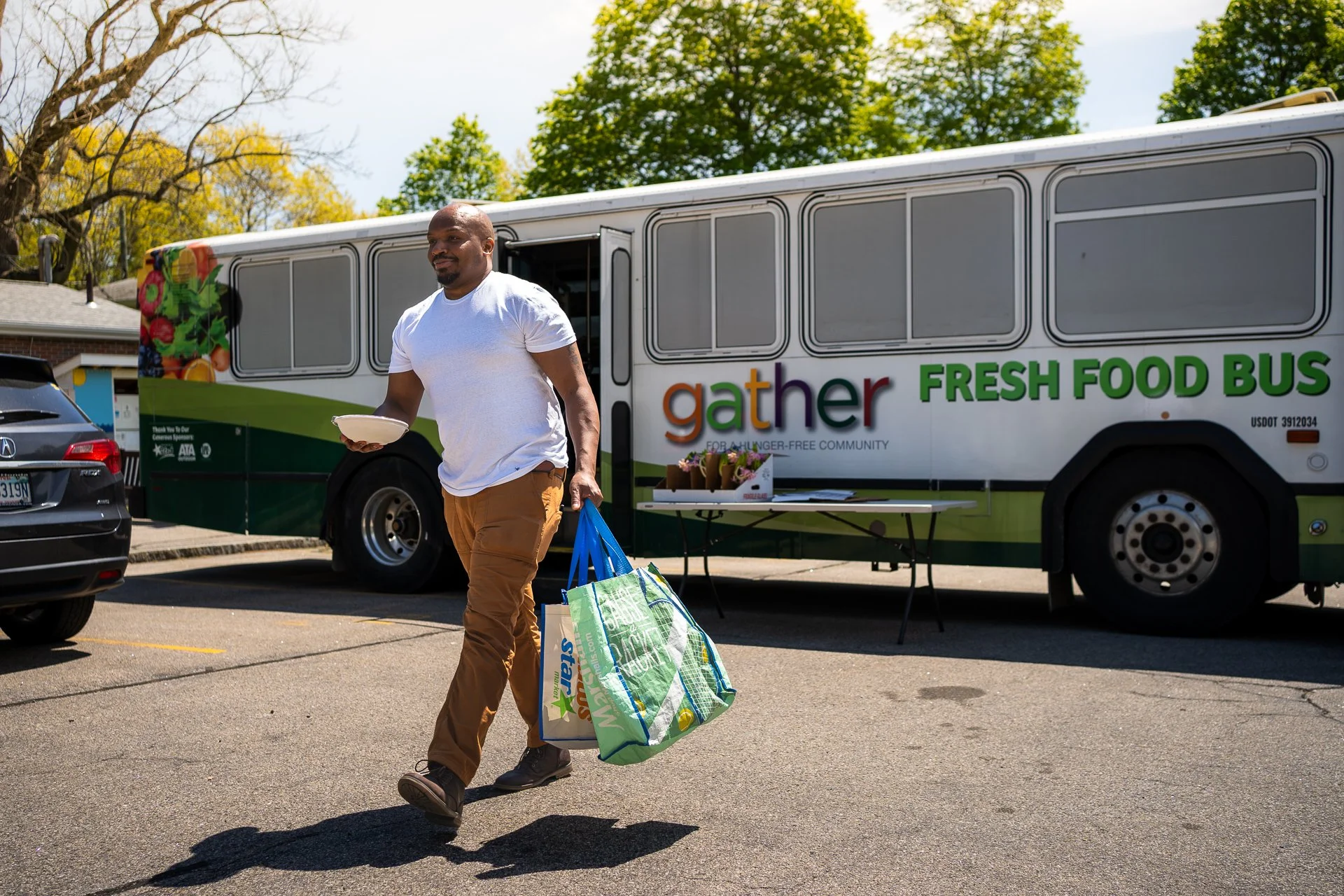 A man walking away from a mobile grocery store with reusable shopping bags, holding a bowl, in a parking lot with trees in the background.