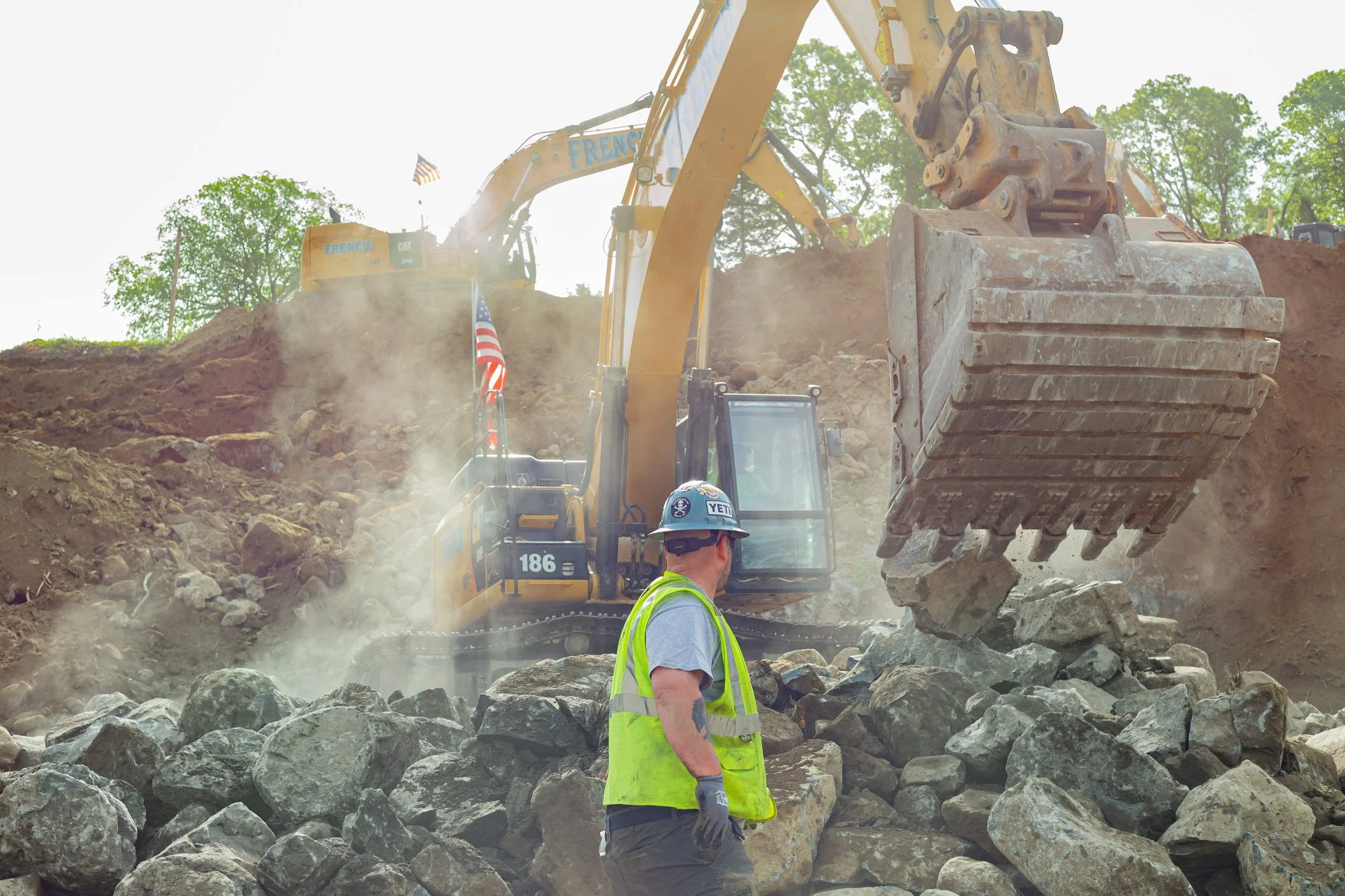 Marketing photo of construction worker in a yellow safety vest and helmet standing among large rocks while excavator with a bucket moves dirt and rocks on a hillside.