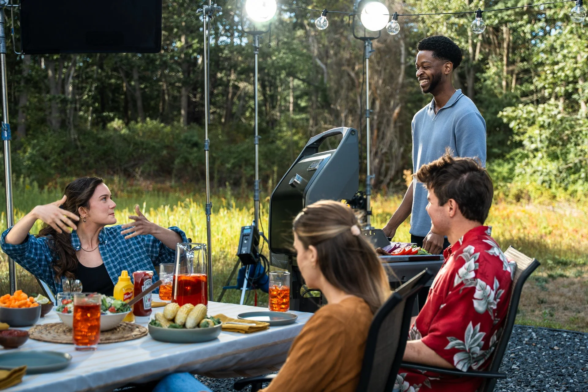 Shooting content of people enjoying a backyard barbecue outdoors with food and drinks on a table, with a filming crew capturing the moment under string lights and trees.